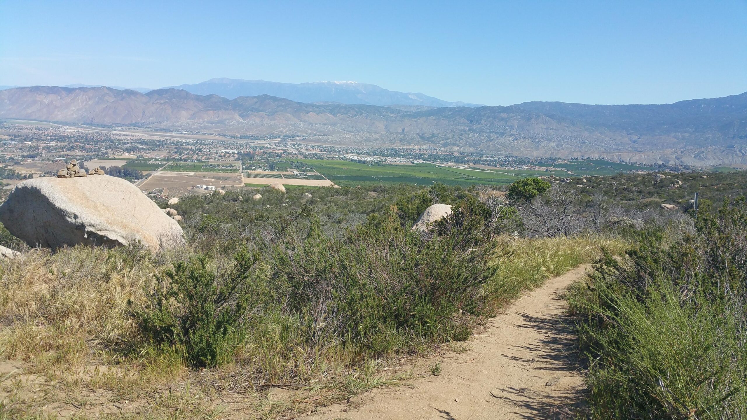 A scenic view from a hillside overlooking a valley, featuring rolling mountains in the background and a patchwork of fields and vineyards below. The foreground includes large boulders and green shrubs, with a dirt path winding through the landscape. Clear blue skies and bright sunlight create a vibrant, natural atmosphere. Simpson Park mountain bike trail.