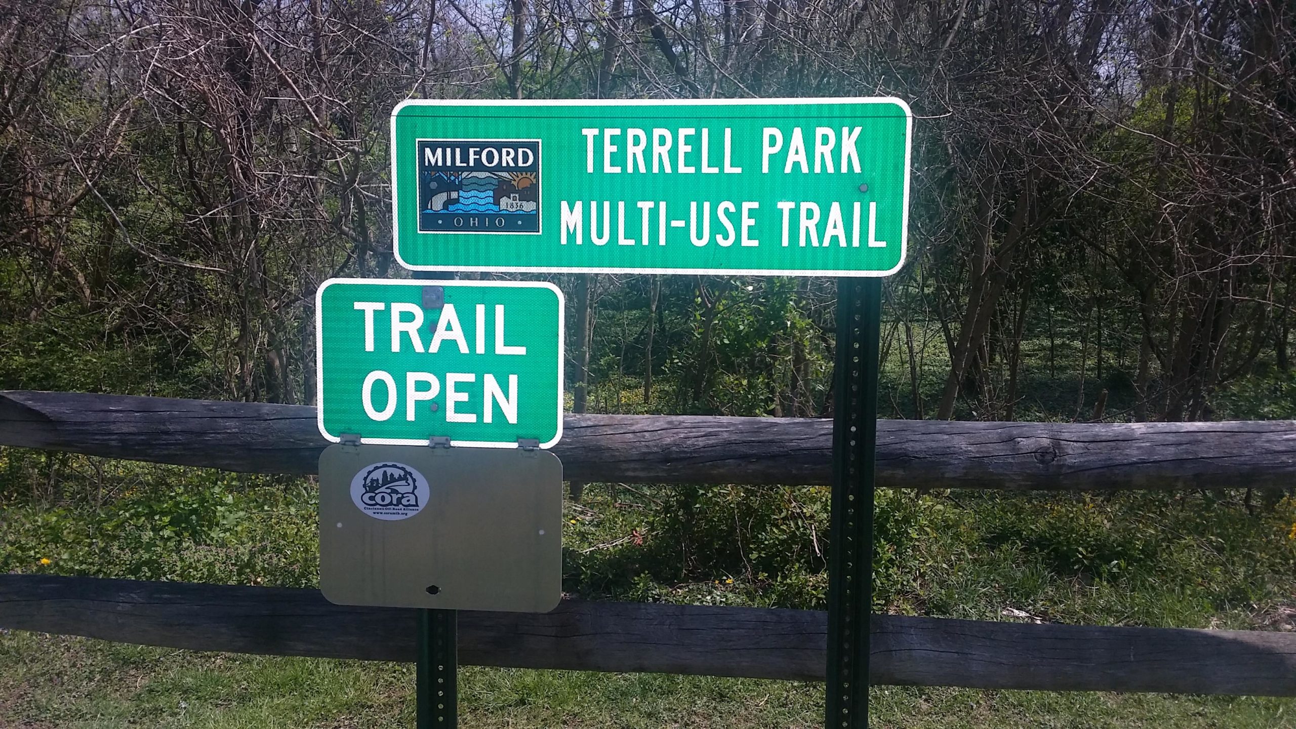 Signage indicating the Terrell Park Multi-Use Trail in Milford, Ohio, with a "Trail Open" notification. The background features a wooded area, and a wooden fence is visible in the foreground. Jim Terrell Park mountain bike trail.