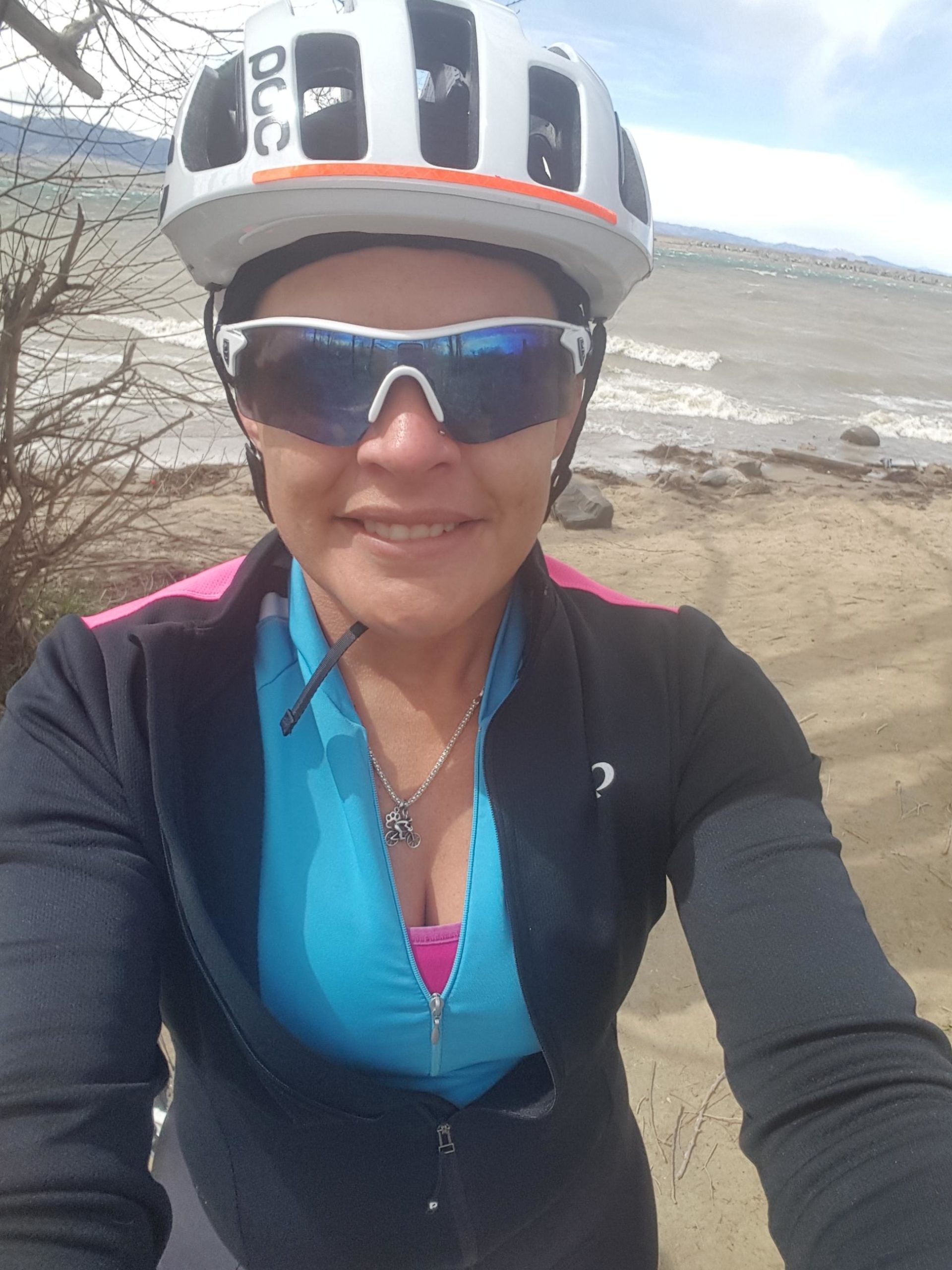 A smiling person wearing a cycling helmet and sunglasses, standing near a sandy beach with water and a distant shoreline in the background. The individual is dressed in a blue cycling jersey with a black jacket over it. Standley Lake mountain bike trail.