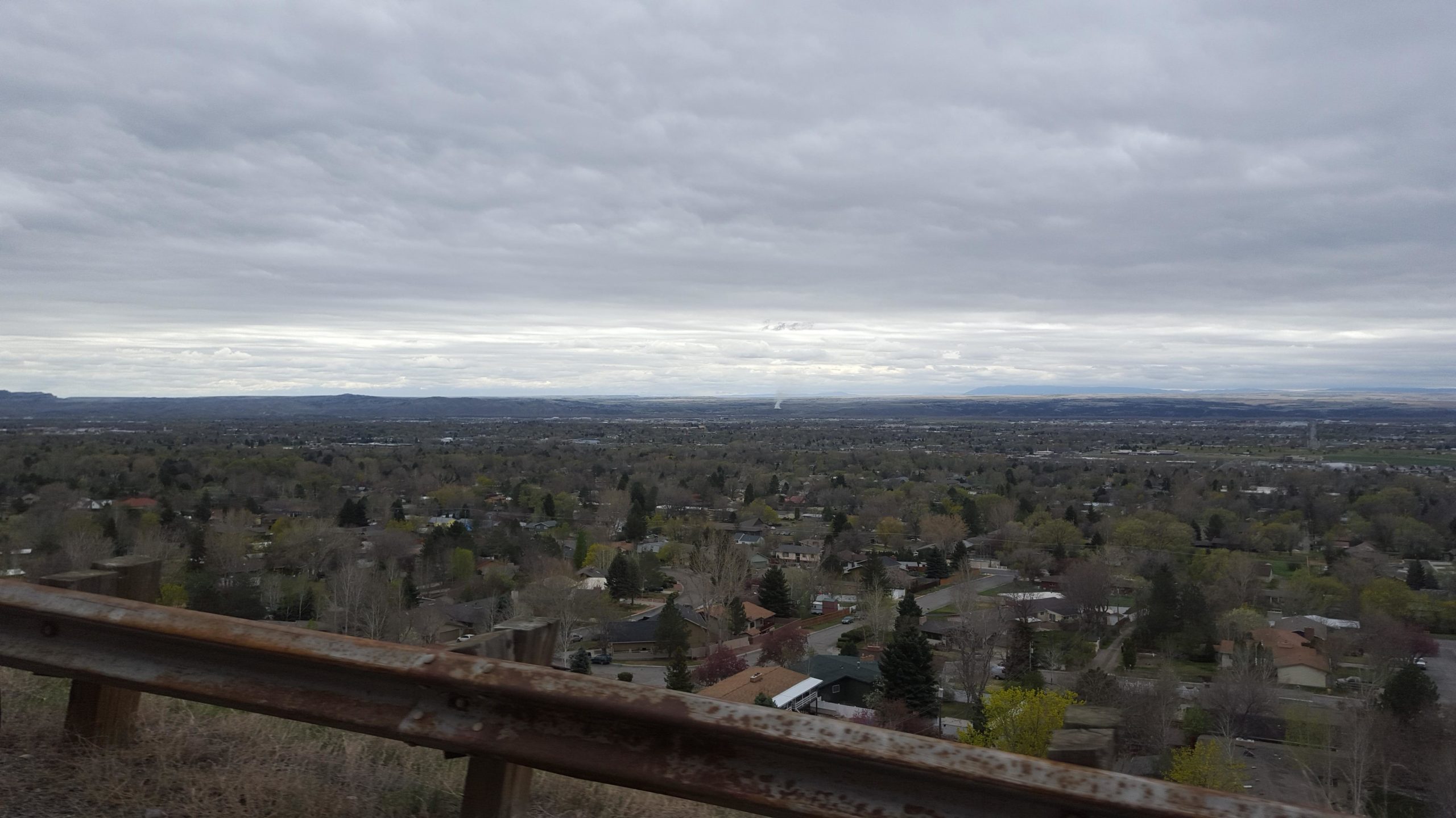 A panoramic view of a valley with wooded areas and residential neighborhoods beneath a cloudy sky, featuring distant mountains and a slight haze in the background. A rusty metal guardrail runs along the edge of the viewpoint. Zimmerman Trail mountain bike trail.