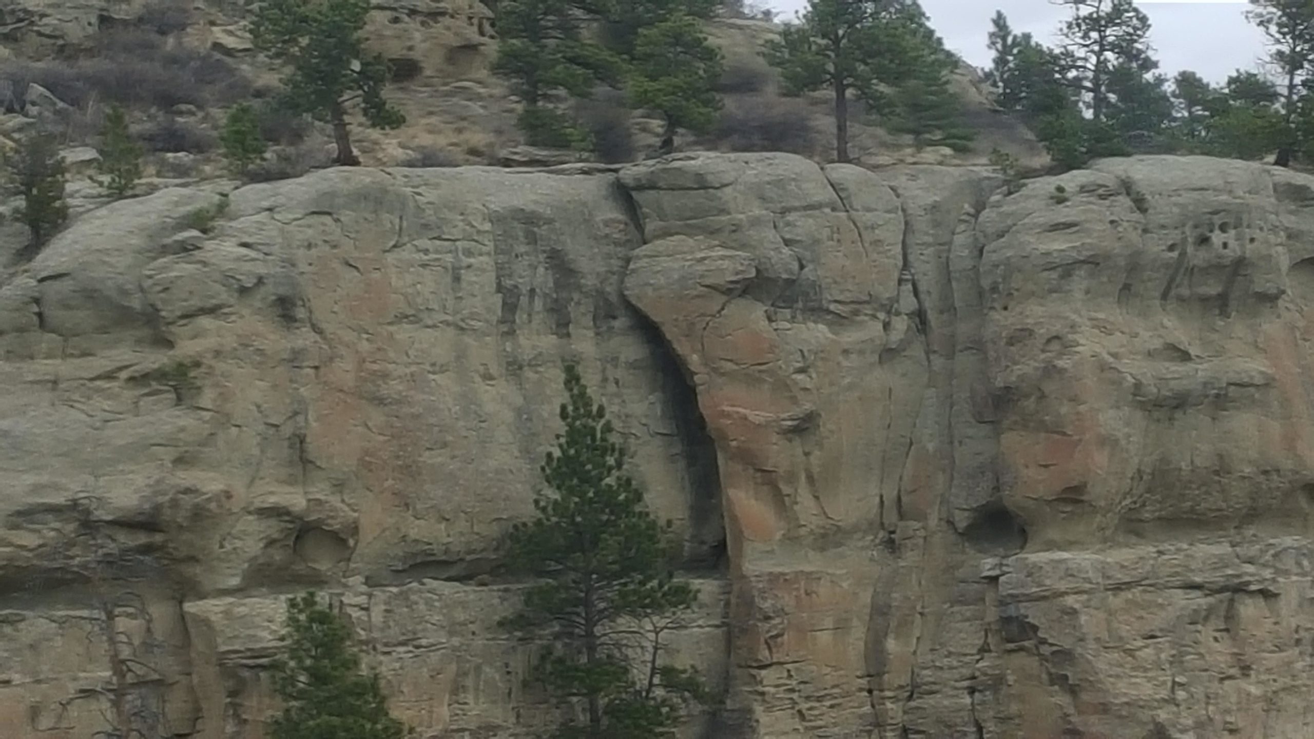 A rocky cliffside featuring rugged stone formations and scattered pine trees at the top, under a cloudy sky. Zimmerman Trail mountain bike trail.