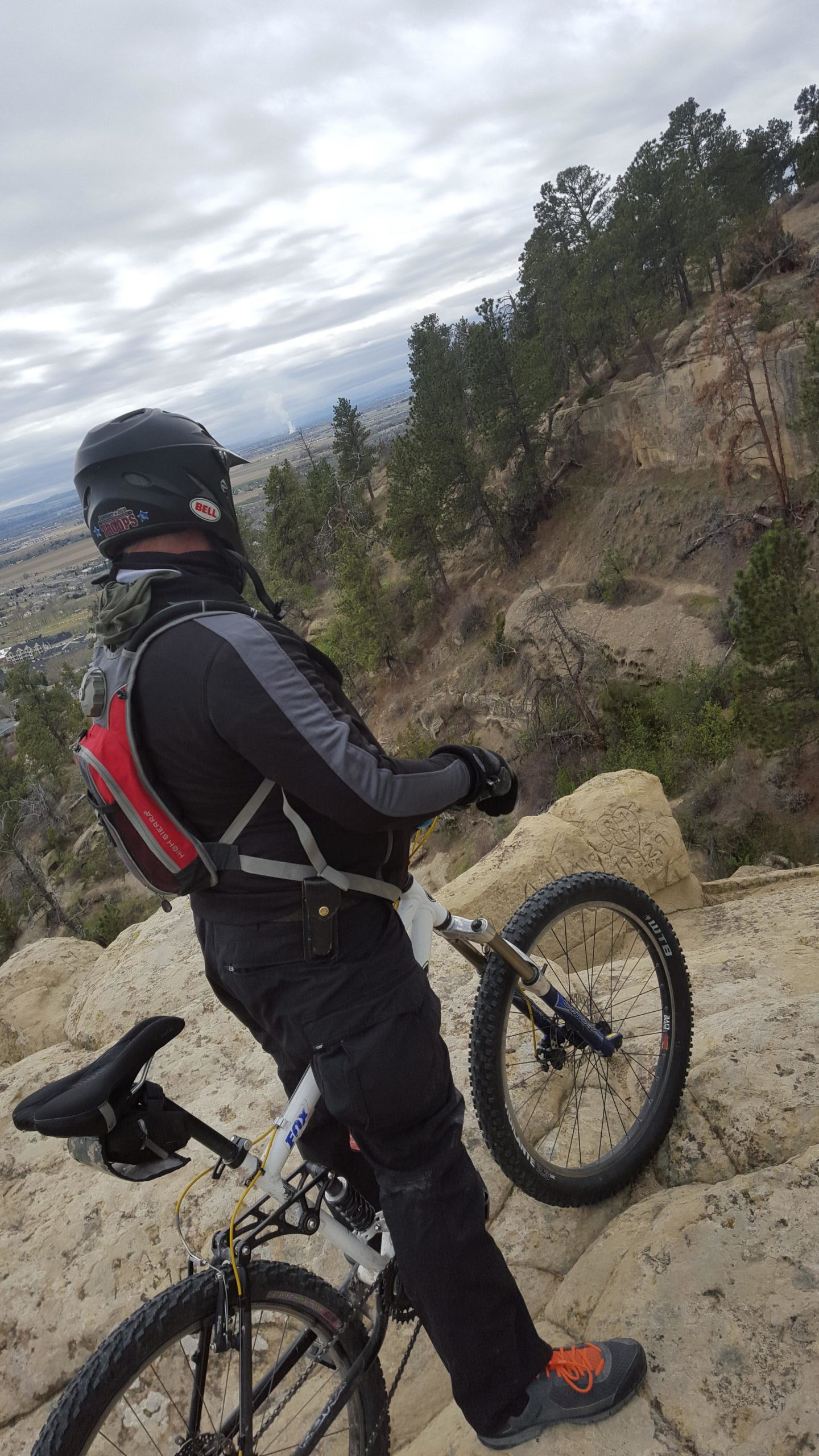 A mountain biker in a helmet and black gear stands next to their bike, overlooking a rocky landscape with trees and a valley in the distance under a cloudy sky. Zimmerman Trail mountain bike trail.