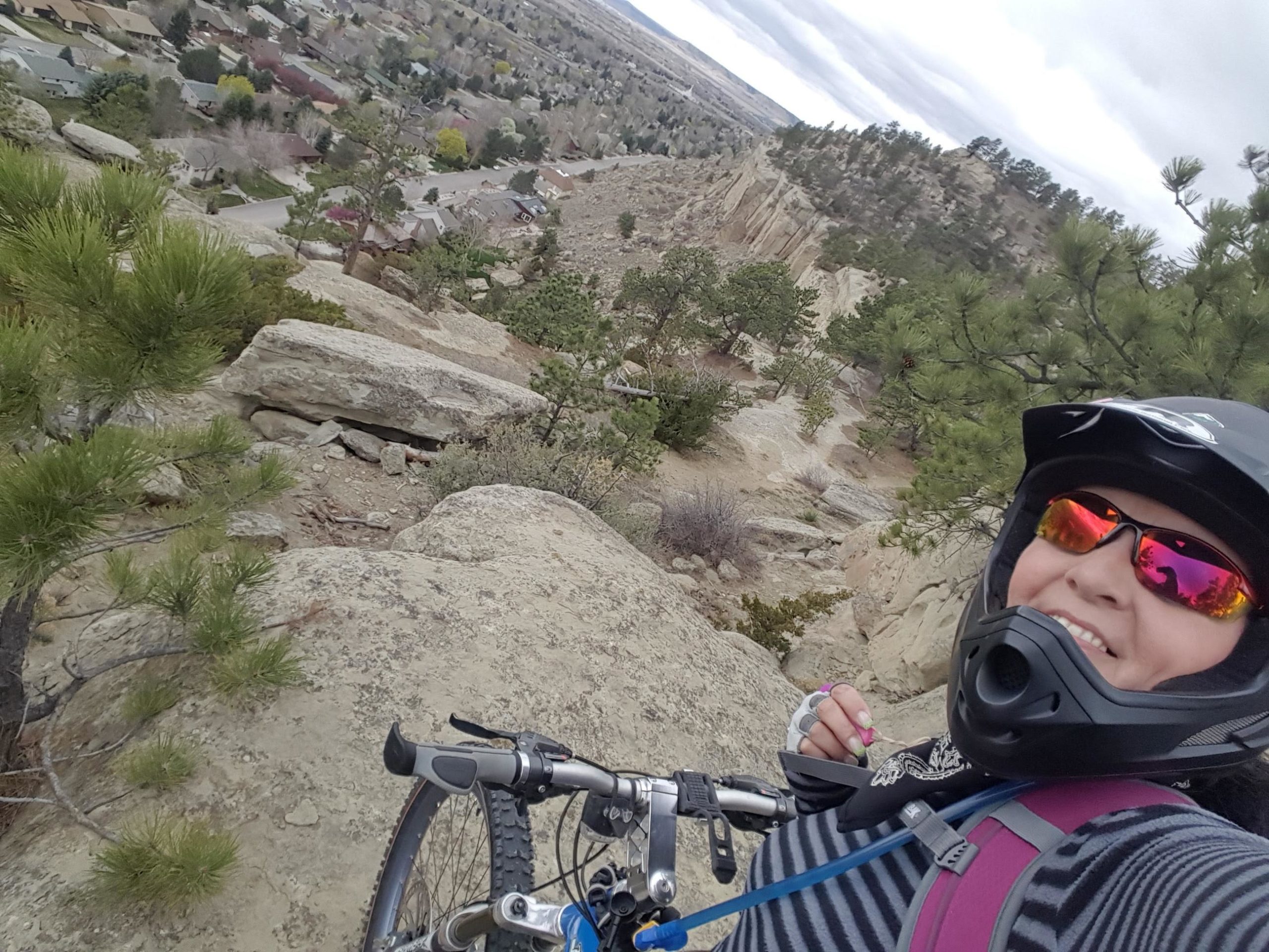 A person wearing a helmet and sunglasses smiles for a selfie while standing next to a mountain bike on rocky terrain, overlooking a valley with scattered homes and trees in the background. The weather appears cloudy. Zimmerman Trail mountain bike trail.