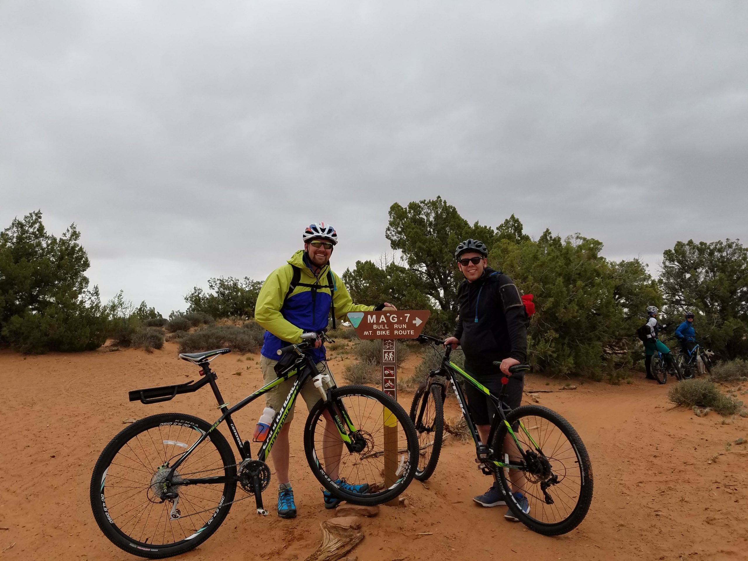 Two mountain bikers posing near a trail sign labeled "MAG-7" in a sandy landscape. The first rider, wearing a green and blue jacket and a helmet, stands next to his bike, while the second rider, dressed in a black jacket and helmet, smiles beside his bike. Lush greenery surrounds them, and other bikers are visible in the background under a cloudy sky. Bull Run mountain bike trail.
