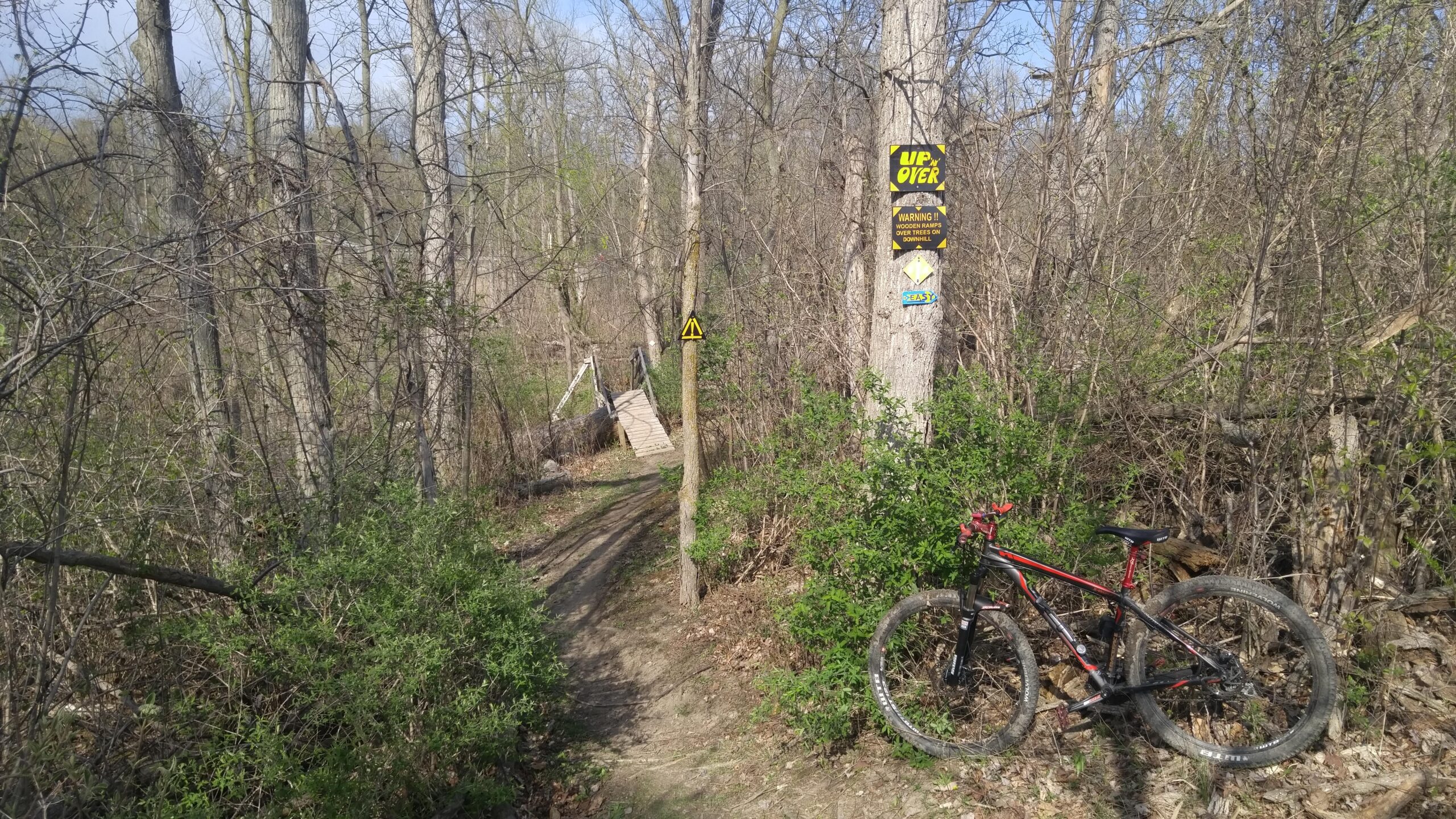 A narrow dirt path through a wooded area, with a mountain bike resting on the ground to the right. A small wooden bridge is visible in the background, accompanied by warning signs on nearby trees indicating trail conditions. The scene is filled with bare trees and green shrubbery, suggesting early spring. Morton-Taylor Trail mountain bike trail.