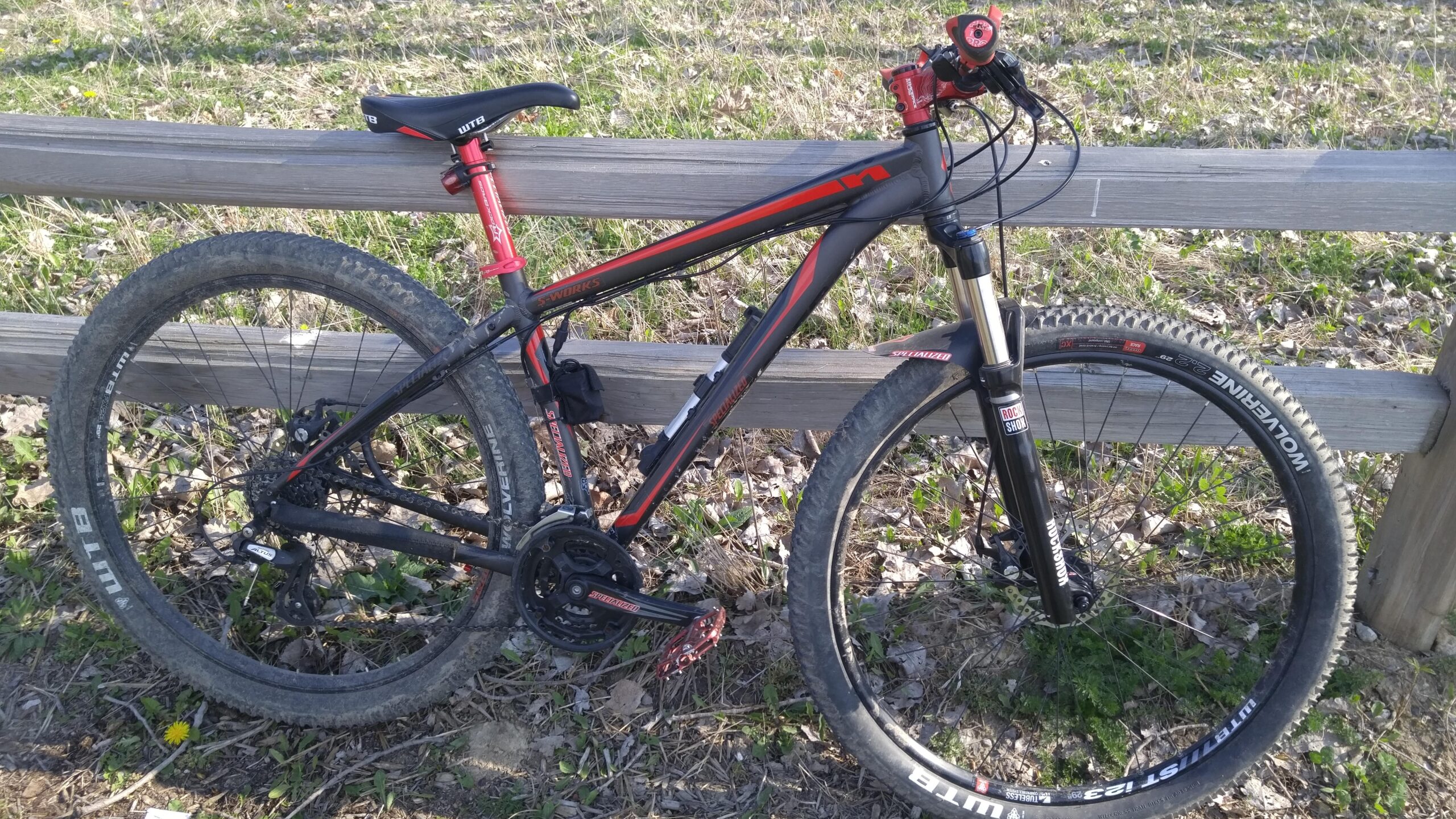 Specialized Hardrock Disc: A mountain bike with a black and red frame, leaning against a wooden fence. The bike features front suspension and has dirt-stained tires, indicating off-road use. In the background, grass and leaves are visible, suggesting a natural outdoor setting.
