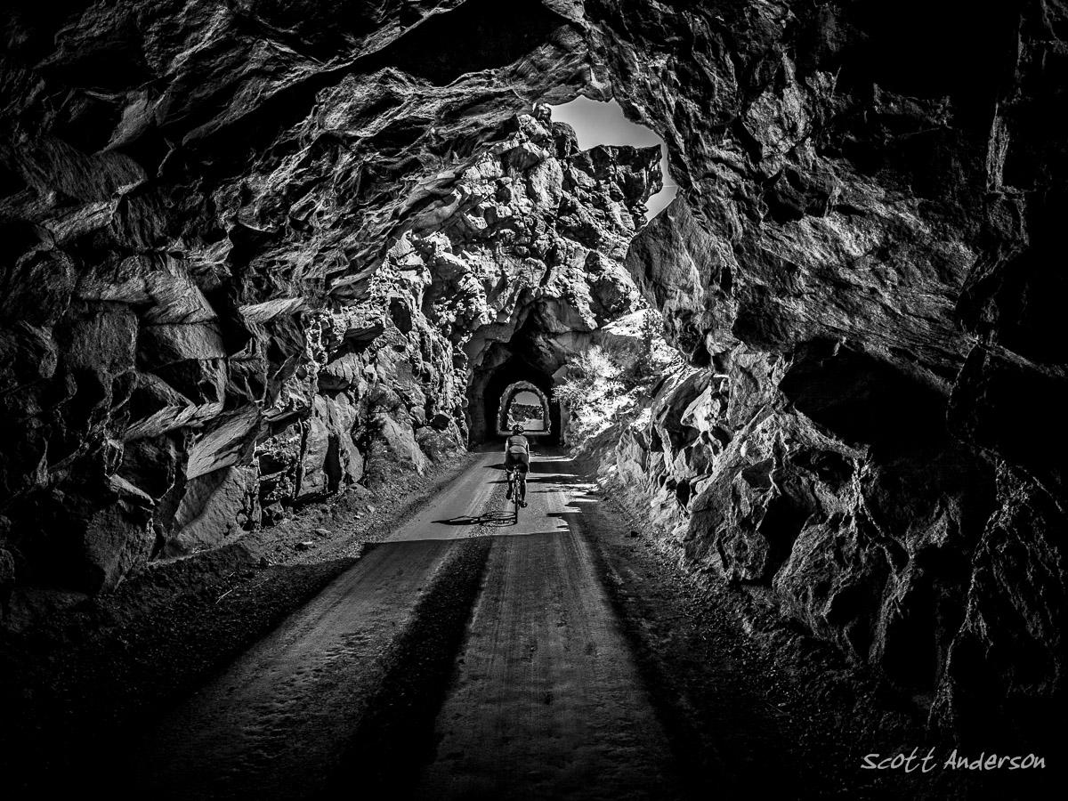 A black and white image of a cyclist riding through a rocky tunnel surrounded by large stone formations. The dirt path leads through the tunnel, with light illuminating the opening in the distance. The texture of the rocks is prominent, creating a dramatic contrast of light and shadow. River Road / #371 mountain bike trail.