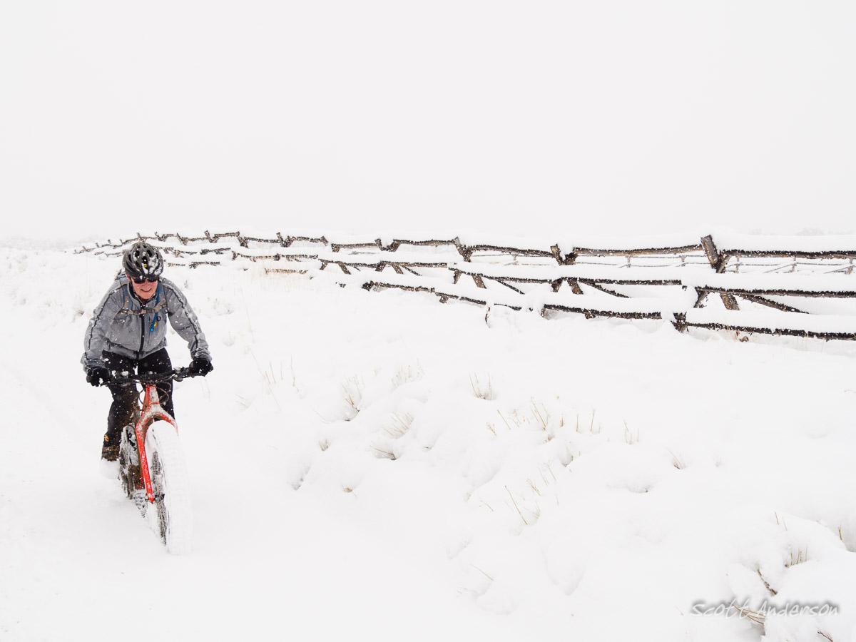 A person riding a fat bike on a snowy path surrounded by a white landscape, with a wooden fence in the background. The scene captures a winter setting with falling snow and a foggy atmosphere, emphasizing the snowy terrain. River Road / #371 mountain bike trail.