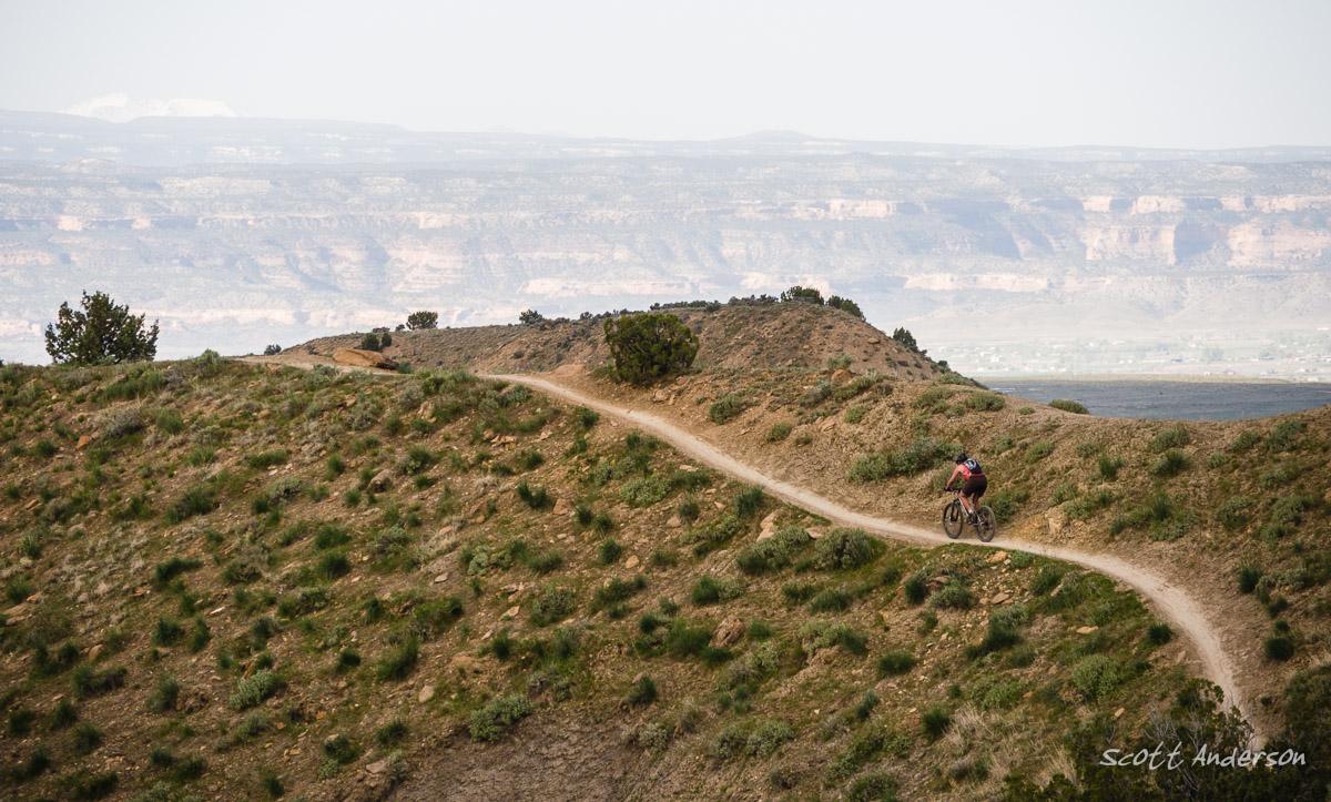 A mountain biker navigating a winding dirt trail on a hillside, surrounded by sparse vegetation and distant rugged cliffs under a hazy sky. Joe's Ridge mountain bike trail.