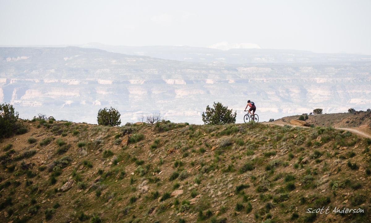 A mountain biker rides along a rugged trail on a hillside, surrounded by sparse vegetation and distant rocky cliffs under a hazy sky. Joe's Ridge mountain bike trail.