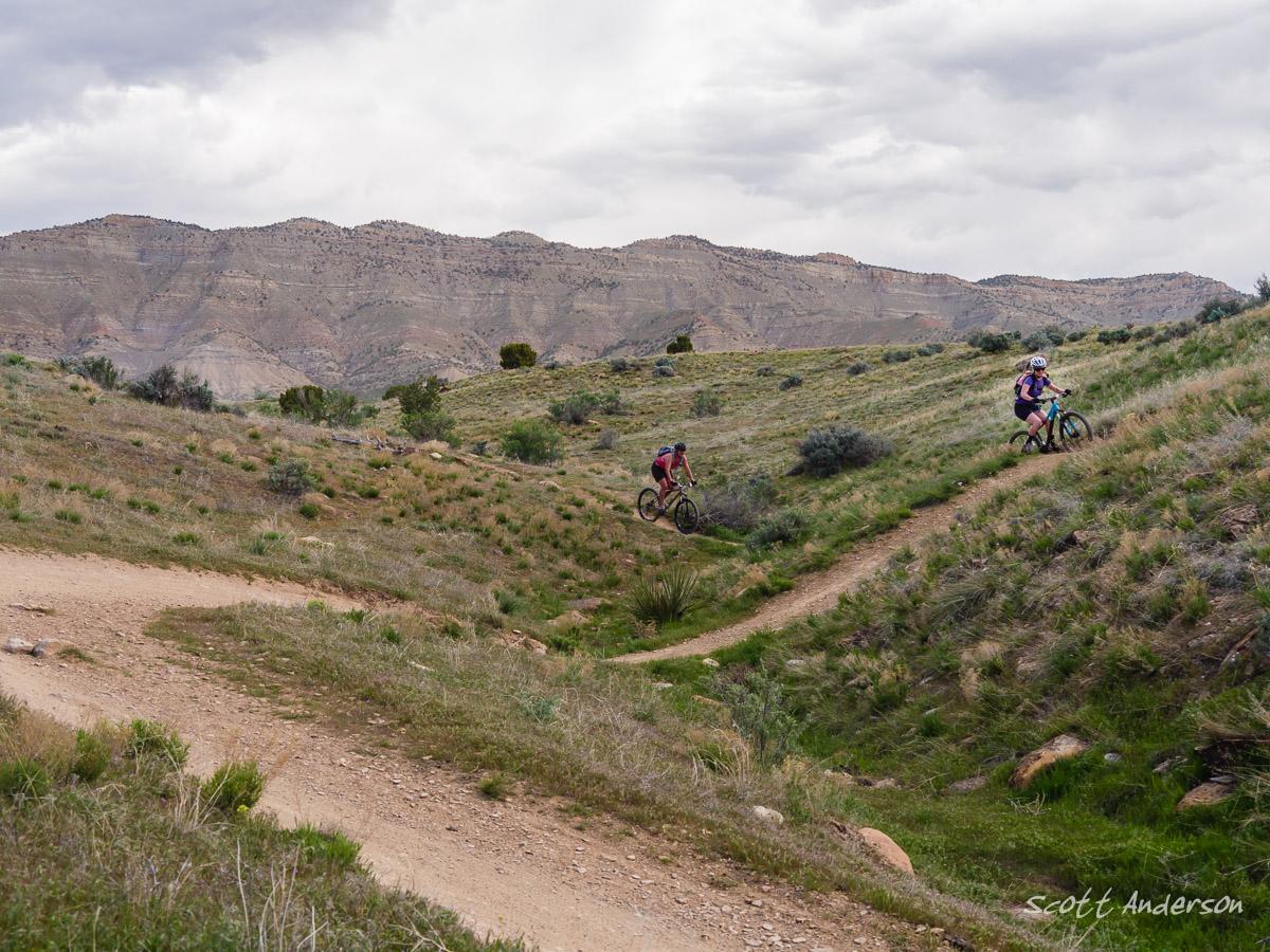 Two mountain bikers navigate a dirt trail through a grassy landscape with rolling hills and sparse vegetation. The background features rugged mountains under a cloudy sky, creating a scenic outdoor environment. Kessel Run mountain bike trail.