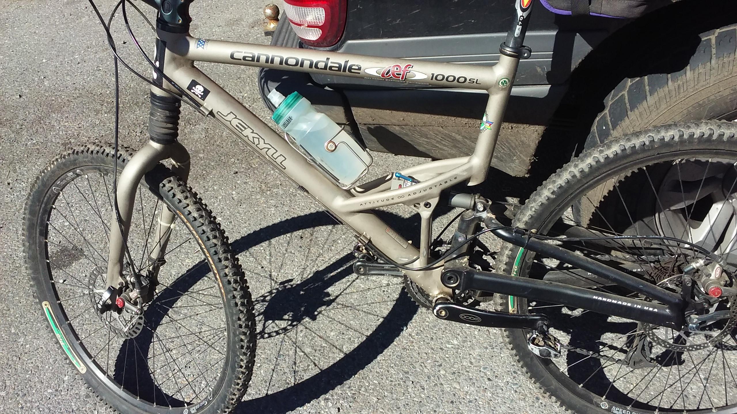 Cannondale Scalpel: Alt text: A close-up view of a Cannondale Jekyll 1000SL mountain bike parked beside a vehicle. The bike features a distinctive frame design, disc brakes on the front wheel, and a water bottle mounted on the frame. Sunlight highlights the bike's metallic color and details, with gravel visible beneath it.
