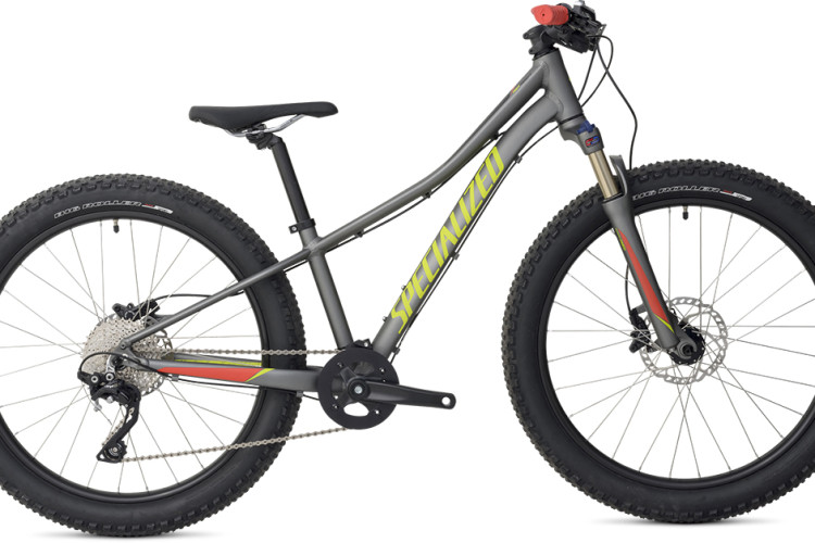 A modern youth mountain bike with a gray frame, featuring a bright yellow and red graphic design. The bike has wide, knobby tires designed for off-road terrain, disc brakes, and a sturdy frame. It is angled to show the side view, highlighting the suspension fork and gearing system.