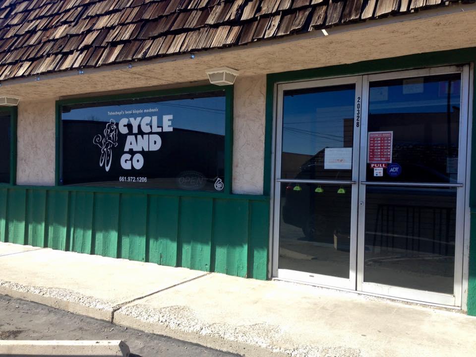 Front facade of "Cycle and Go," a bicycle repair shop with large windows displaying the business name and contact number. The building features a green bottom half and a brown shingled roof, indicating a welcoming atmosphere for cyclists. A small sign on the door shows it is open for business.