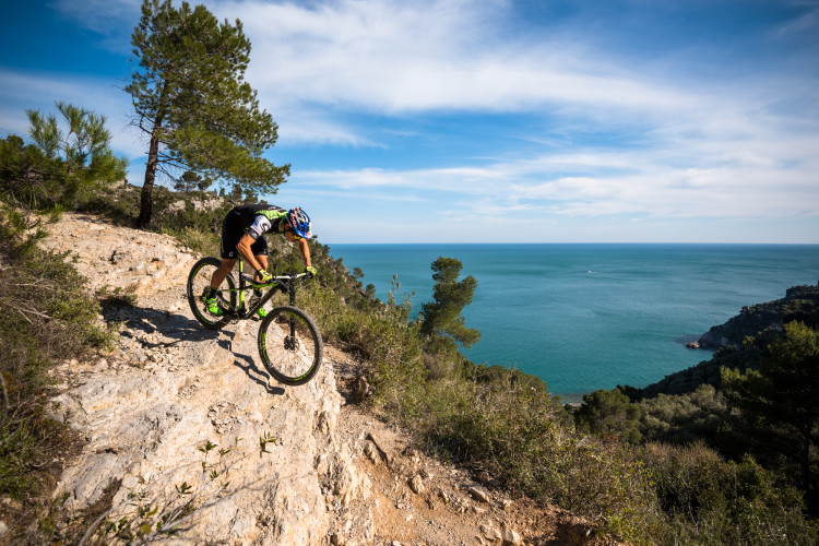 A mountain biker navigating a rocky trail along a coastal cliff, with the ocean and blue sky in the background. Green trees and vegetation surround the path, creating a scenic outdoor setting.