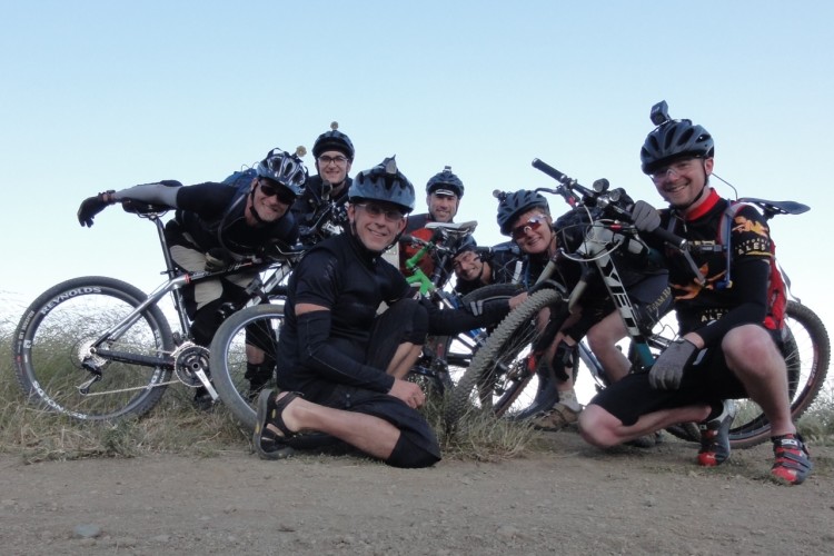 A group of six mountain bikers, wearing helmets and cycling gear, pose together on a dirt trail. They are smiling and crouched around their bicycles, showcasing a sense of camaraderie and adventure. The background features tall grass and a clear blue sky.