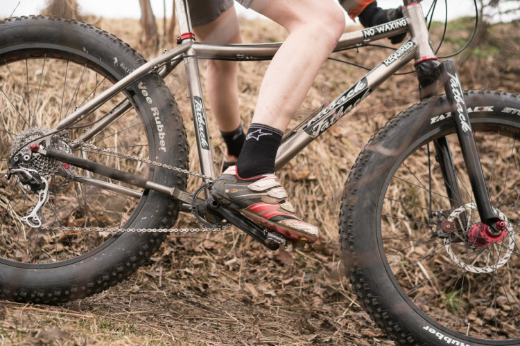 A close-up view of a mountain biker's legs and a fat bike, showcasing the bike's thick tires and chain as the rider pedals on rough terrain covered with grass and leaves.