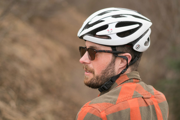 A man wearing a white cycling helmet and dark sunglasses stands outdoors, looking to the side. He has a beard and is dressed in a plaid shirt, with a blurred natural background of earthy tones. The image captures a moment of contemplation or focus while enjoying an outdoor activity.