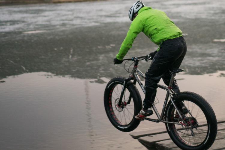 A cyclist in a bright green jacket and helmet rides a fat bike, preparing to jump off a wooden ramp over a frozen body of water. The scene captures the adventurous spirit of winter cycling.