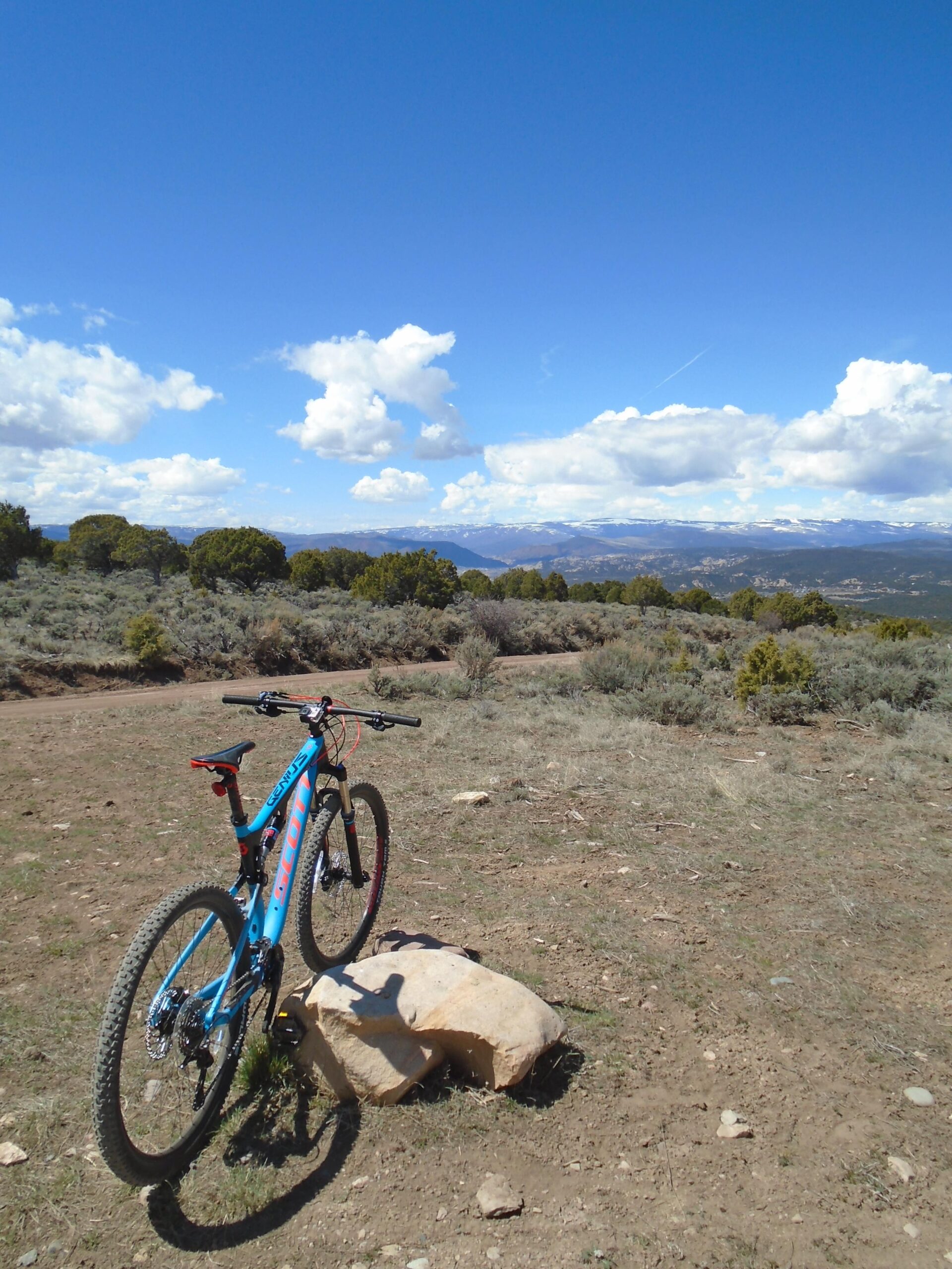 Scott Genius LT 720: A blue mountain bike rests against a large rock on a dirt trail, surrounded by shrubs and sparse vegetation. In the background, there are distant mountains under a clear blue sky with fluffy white clouds.
