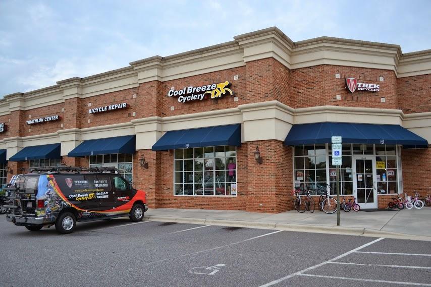 Image of a brick storefront featuring "Cool Breeze Cyclery" and "Trek Bicycles" signs. The shop has large windows displaying bicycles and related accessories. A branded van is parked in front, and there are several bicycles outside. The scene is set in a suburban shopping area. Blue awnings cover the windows, and the sky is slightly overcast.