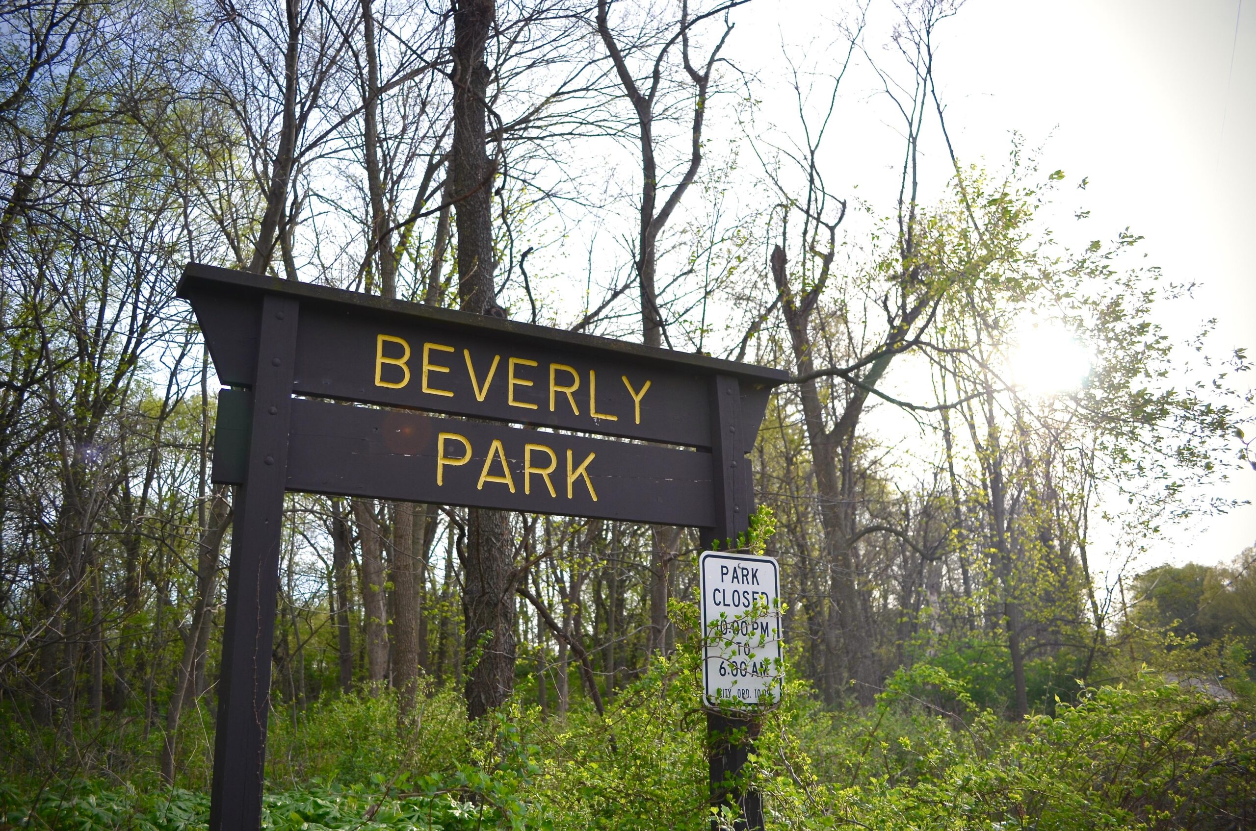 A wooden sign reading "Beverly Park" stands amidst a green, wooded area, with trees and foliage surrounding it. A second sign below indicates park hours, stating "Park Closed 9:00 PM to 6:00 AM." The sun shines softly in the background, illuminating the scene. Beverly Park mountain bike trail.