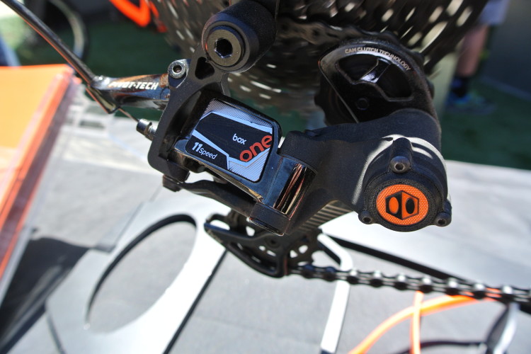 Close-up view of a bicycle derailleur, specifically a Box One 11-speed model, showing details of its construction, including the mechanism and cable attachment. The derailleur is mounted on a bike, with a chain visible in the background.