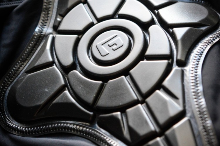 Close-up of a protective gear pad featuring a shiny, molded design. The pad is attached to black fabric with visible stitching. The intricate shape suggests it is designed for impact resistance, likely intended for sports or safety applications.