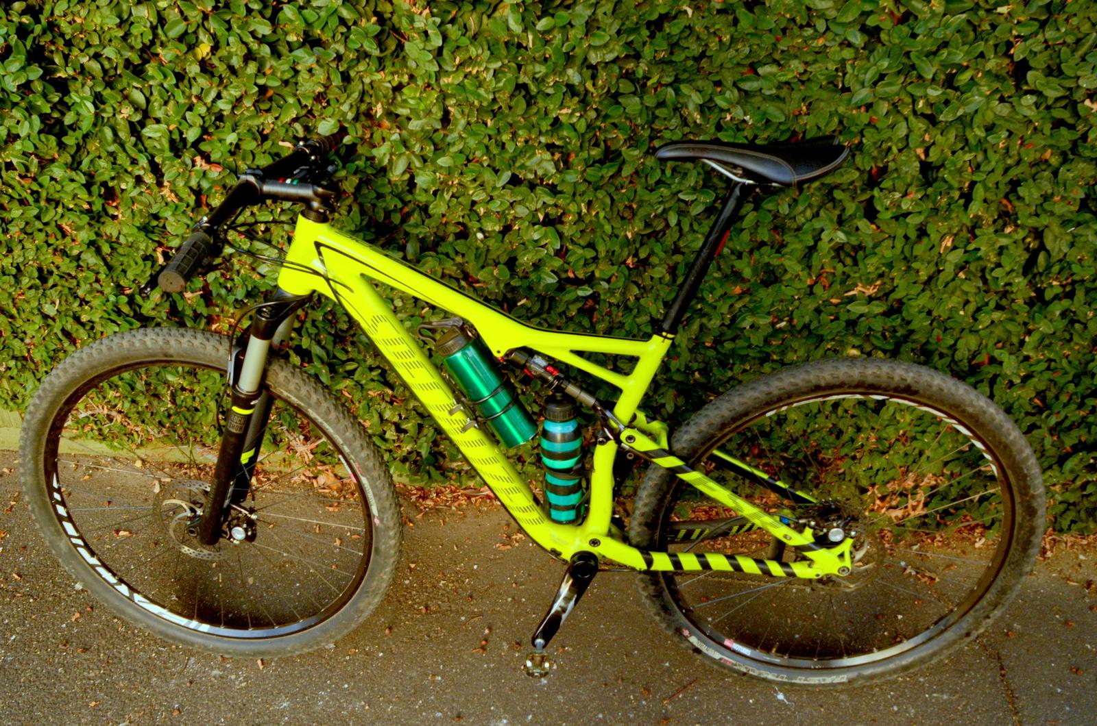 Specialized Epic Comp 29er: A bright yellow mountain bike with a sleek design is positioned against a green shrub background. The bike features two water bottles mounted on the frame, one green and one striped, and has wide tires suited for off-road biking. The handlebars are equipped with grips, and the saddle is black, complementing the vibrant frame color.