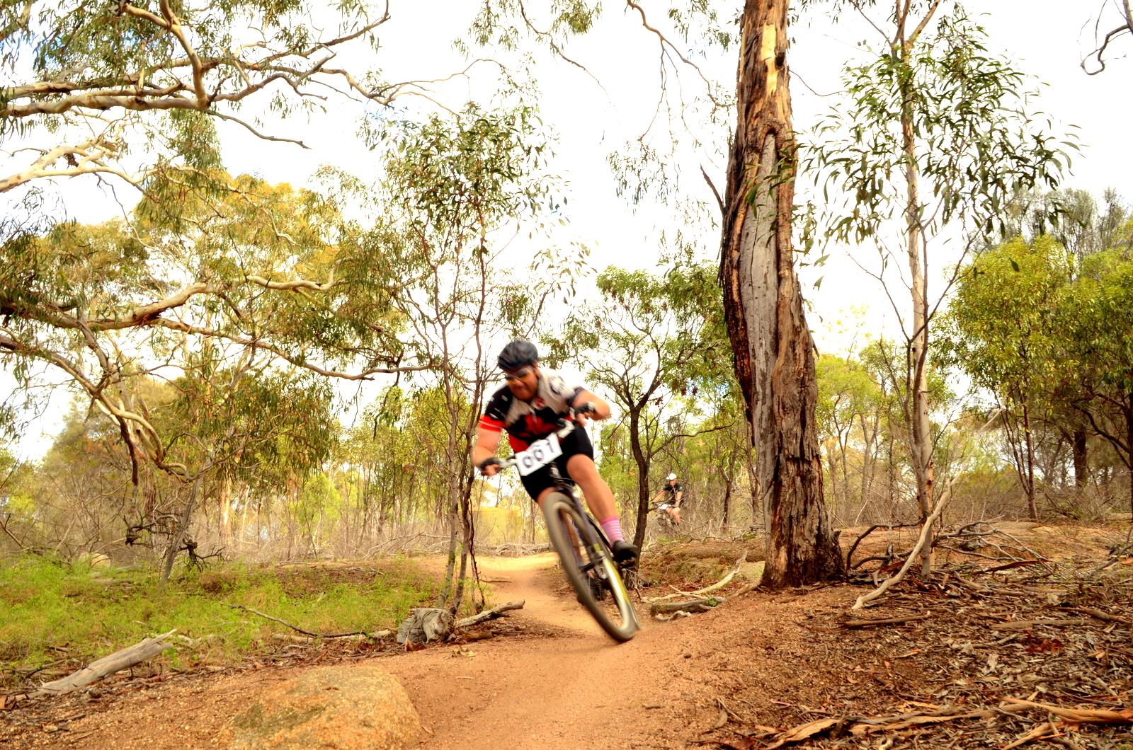 Mountain biker navigating a dirt trail through a wooded area, with trees and bushes surrounding the path. The cyclist is in action, leaning into a turn, and is wearing a helmet and cycling gear. A second biker is visible in the background. You Yangs Regional Park mountain bike trail.