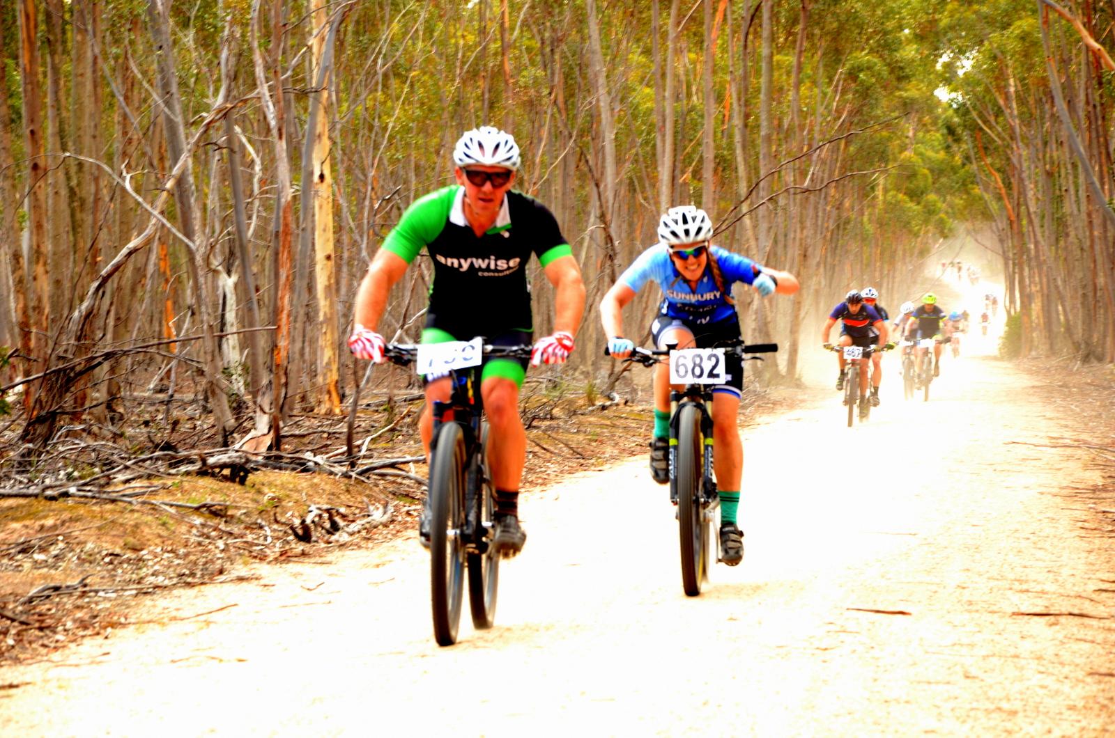 A group of mountain bikers competing on a dusty trail surrounded by tall eucalyptus trees. Two riders, one in a black and green jersey and the other in a blue jersey, are prominently leading the pack, with visible race numbers on their bikes. The background shows additional cyclists moving along the trail, and dust is kicked up as they ride. You Yangs Regional Park mountain bike trail.