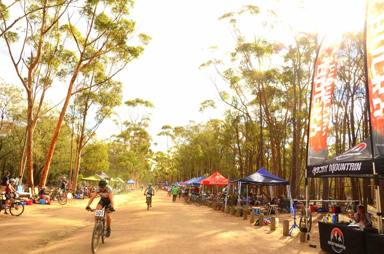 A dirt path lined with tall trees, where cyclists pass by during a mountain biking event. Tents from various sponsors are set up along the side, with people sitting and preparing for the race. The atmosphere is lively and outdoorsy, surrounded by nature. You Yangs Regional Park mountain bike trail.