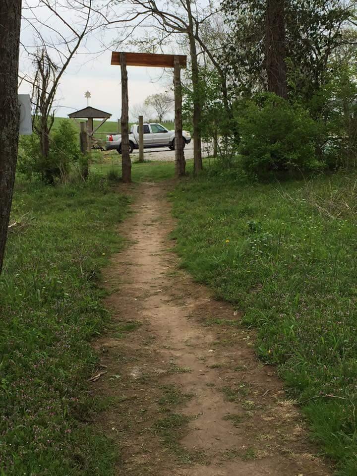 A dirt path leading through greenery, with a wooden archway at the entrance. In the background, there is a parking area with a vehicle visible. The scene is set against a cloudy sky, suggesting a peaceful outdoor environment. Knucklehead mountain bike trail.