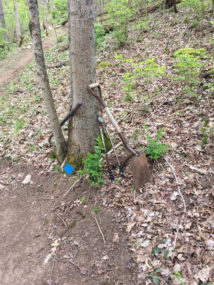 Gardening tools, including a shovel, lopper, and a small blue marker, leaning against a tree in a wooded area covered with fallen leaves and small green plants. A dirt path is visible in the background. Knucklehead mountain bike trail.