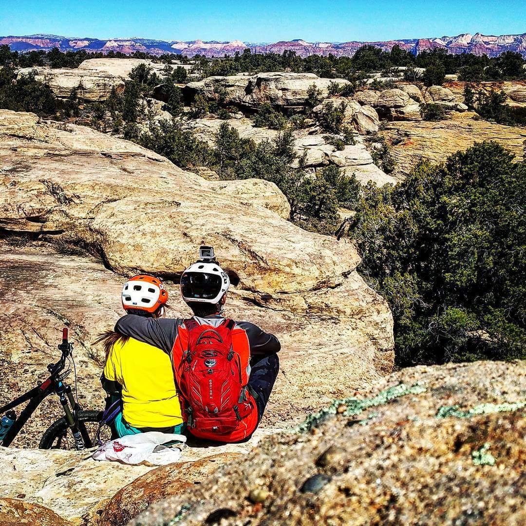 A couple sits on a rocky outcrop, overlooking a scenic landscape filled with trees and distant mountains. They are both wearing helmets, with one wearing a bright yellow shirt and the other in a black shirt with a red backpack. A mountain bike is resting nearby as they enjoy the view together. Gooseberry Mesa mountain bike trail.