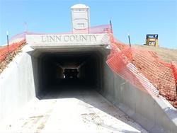 A concrete tunnel entrance with the inscription "Linn County" at the top, surrounded by orange safety fencing and construction signs. The tunnel leads into a shaded area with visible light at the end. Clear blue sky is visible above the tunnel. Grant Wood Trail mountain bike trail.