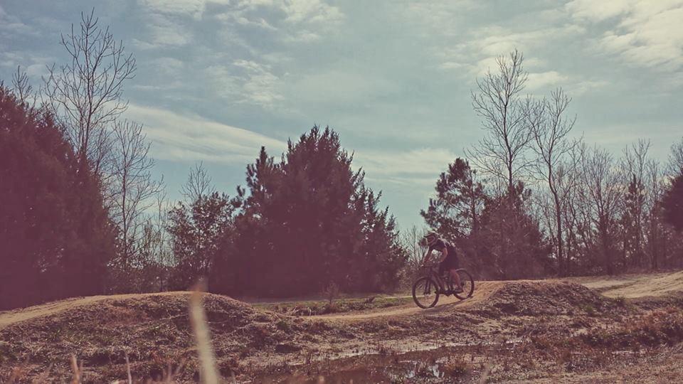 A cyclist riding on a dirt path through a wooded area, with trees in the background and a partly cloudy sky overhead. Horry County Bike Run Park mountain bike trail.