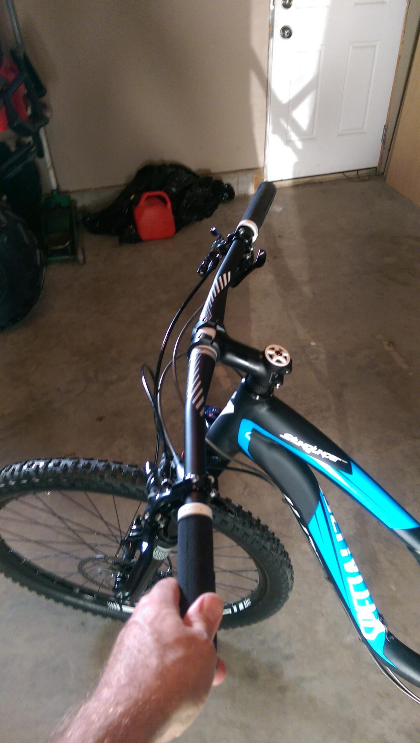 Specialized Stumpjumper FSR Comp 29er: A close-up view of a person's hand gripping the handlebars of a mountain bike. The bike features a black and blue frame, with textured rubber grips. In the background, there is a garage setting with tools and a red fuel canister.