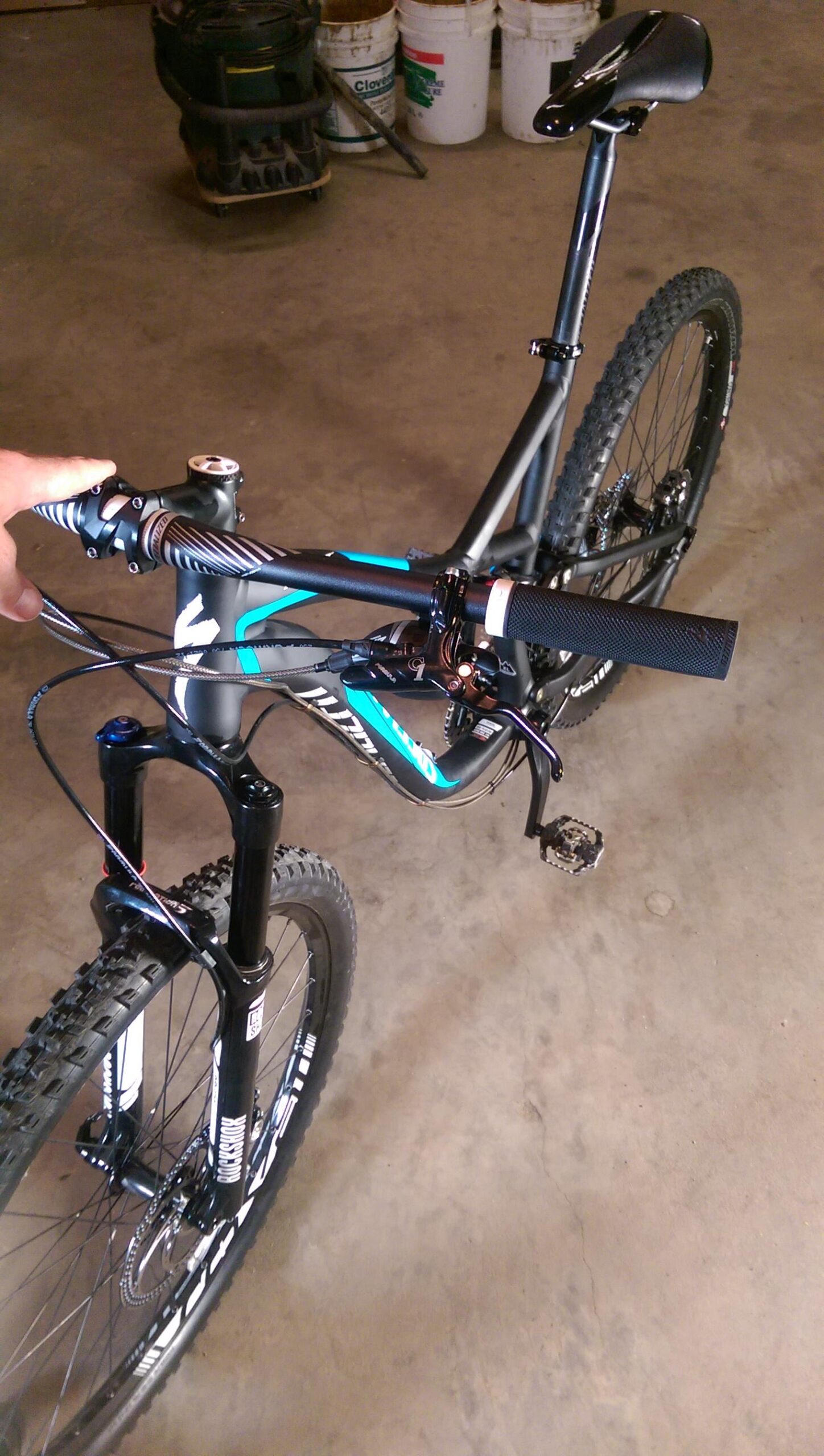 Specialized Stumpjumper FSR Comp 29er: Image of a mountain bike in a garage setting, showing the handlebars, front suspension, and grip. The bike features a sleek black and turquoise design, with a visible front wheel and pedals, and a neutral concrete floor in the background.