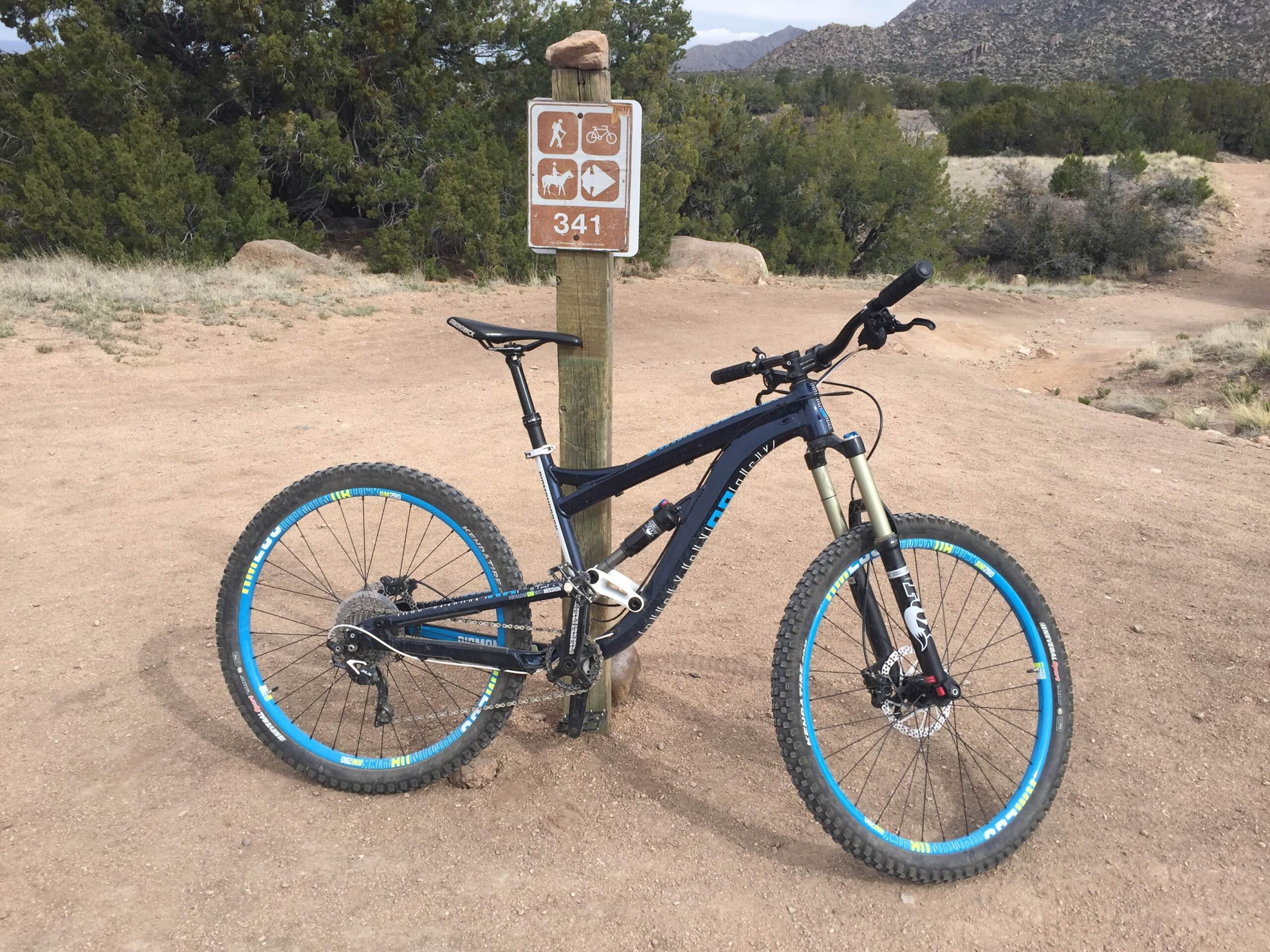 Diamondback Mission 1 27.5: A mountain bike leaning against a trail sign that indicates various activities including hiking and wildlife. The scene is set in a natural outdoor environment with dirt paths and vegetation, surrounded by rocky hills in the background.