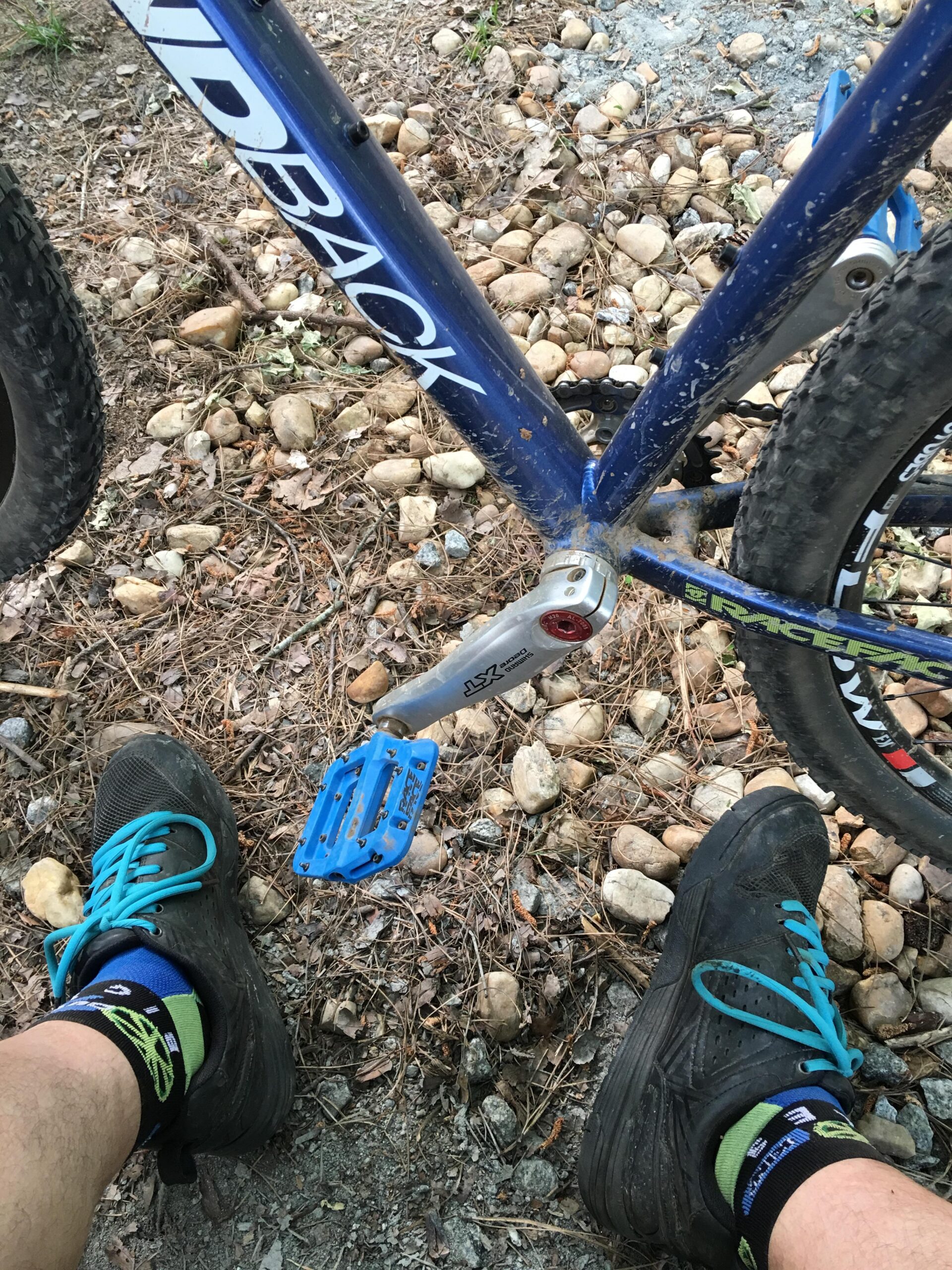 A close-up view of a mountain bike's blue frame and black pedal, with a person's legs and feet visible at the bottom wearing black shoes and colorful socks. The ground is covered in small stones and pine needles, indicating an outdoor setting. Allatoona Creek Park mountain bike trail.