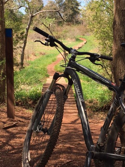 A mountain bike parked next to a trail sign, with a winding dirt path ahead surrounded by greenery and trees. The path features a wooden bridge section, leading into a scenic outdoor setting. Six Mile Run mountain bike trail.