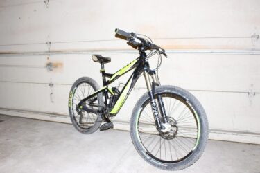 Cannondale Jekyll 3: A black and green mountain bike is leaning against a garage wall. The bike features a sturdy frame, wide tires with visible tread, and front suspension forks. The background shows a plain garage door with a slight texture, emphasizing the bike's design.
