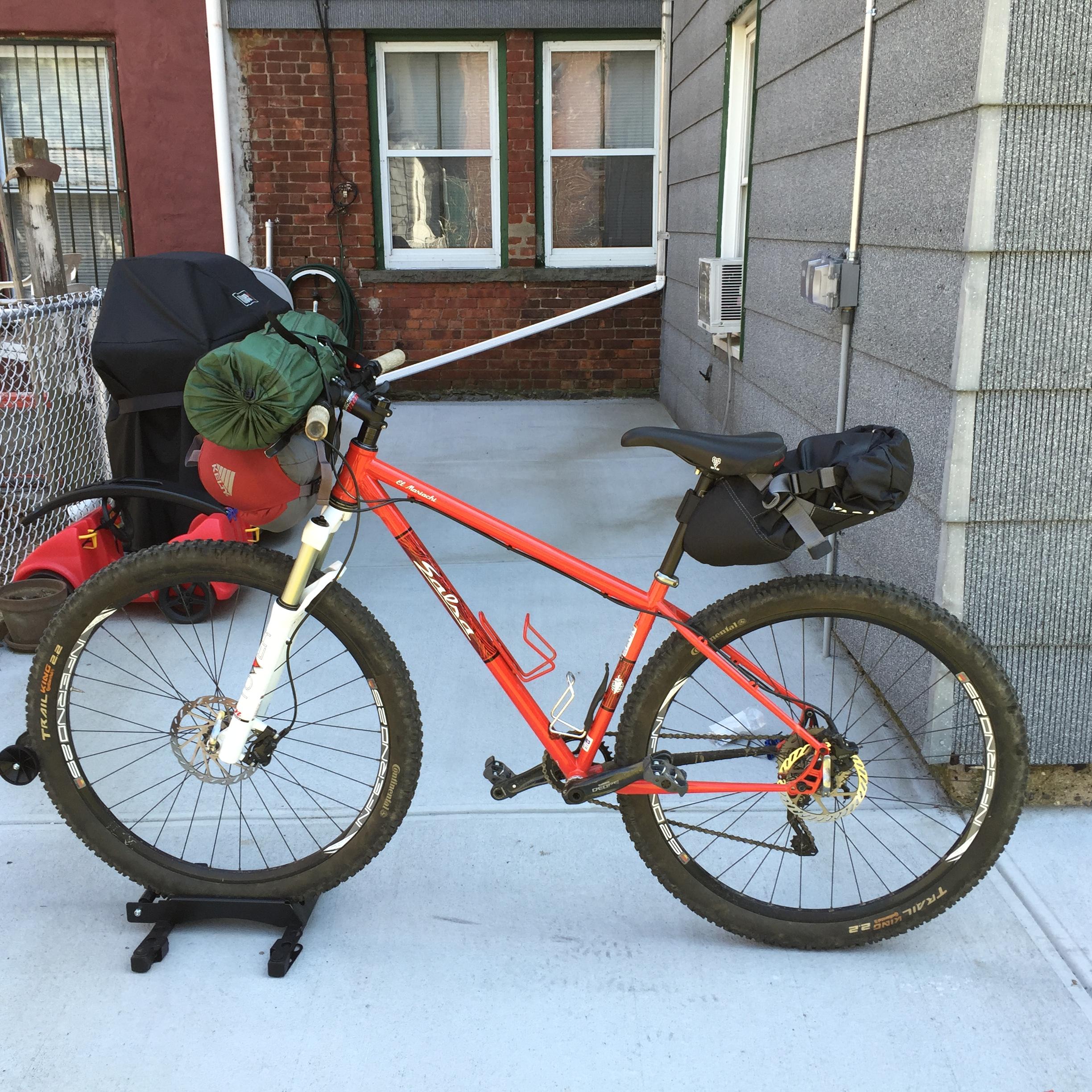 Salsa El Mariachi: A red mountain bike parked on a concrete surface, equipped with various bags and gear on the handlebars and seat, next to a brick wall with windows in the background and an air conditioning unit on the side.