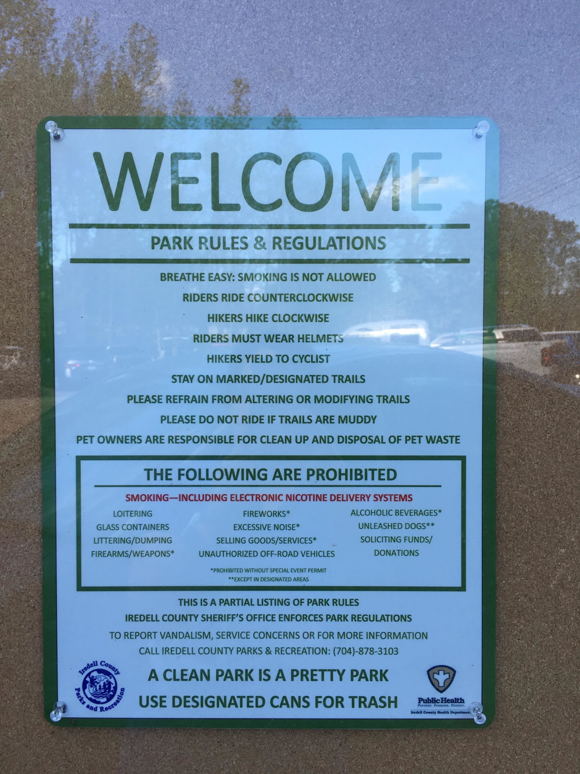 A sign displaying park rules and regulations, including a welcome message and guidelines for visitors. It states that smoking is not allowed, outlines how cyclists and hikers should share the trails, and lists prohibited activities such as loitering, fireworks, and littering. Contact information for local authorities and a reminder to use designated trash cans are also provided. Bigleaf Slopes Park mountain bike trail.