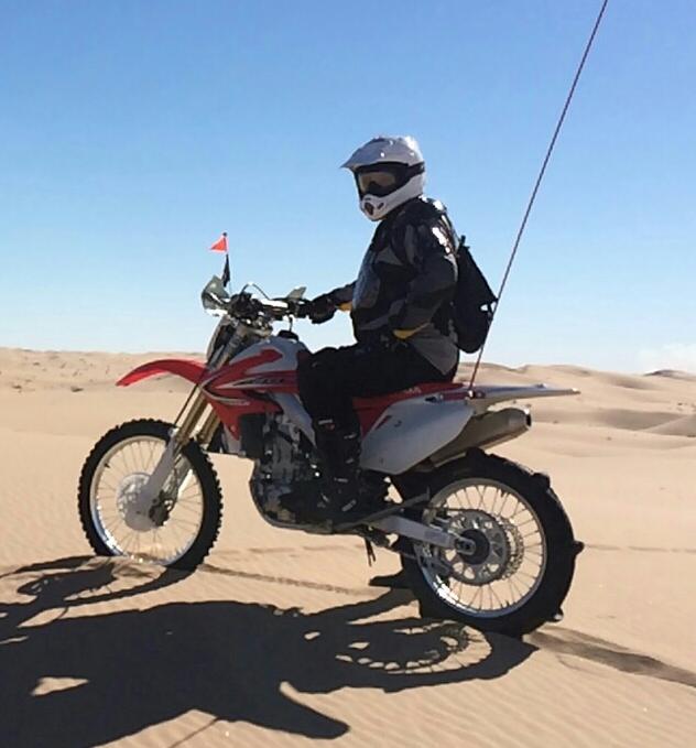 Giant XTC Composite 29er 1: A motorcyclist wearing a helmet and protective gear is riding a red and silver dirt bike on a sandy dune under a clear blue sky. A small flag is attached to the bike, and the landscape features rolling sand hills in the background.