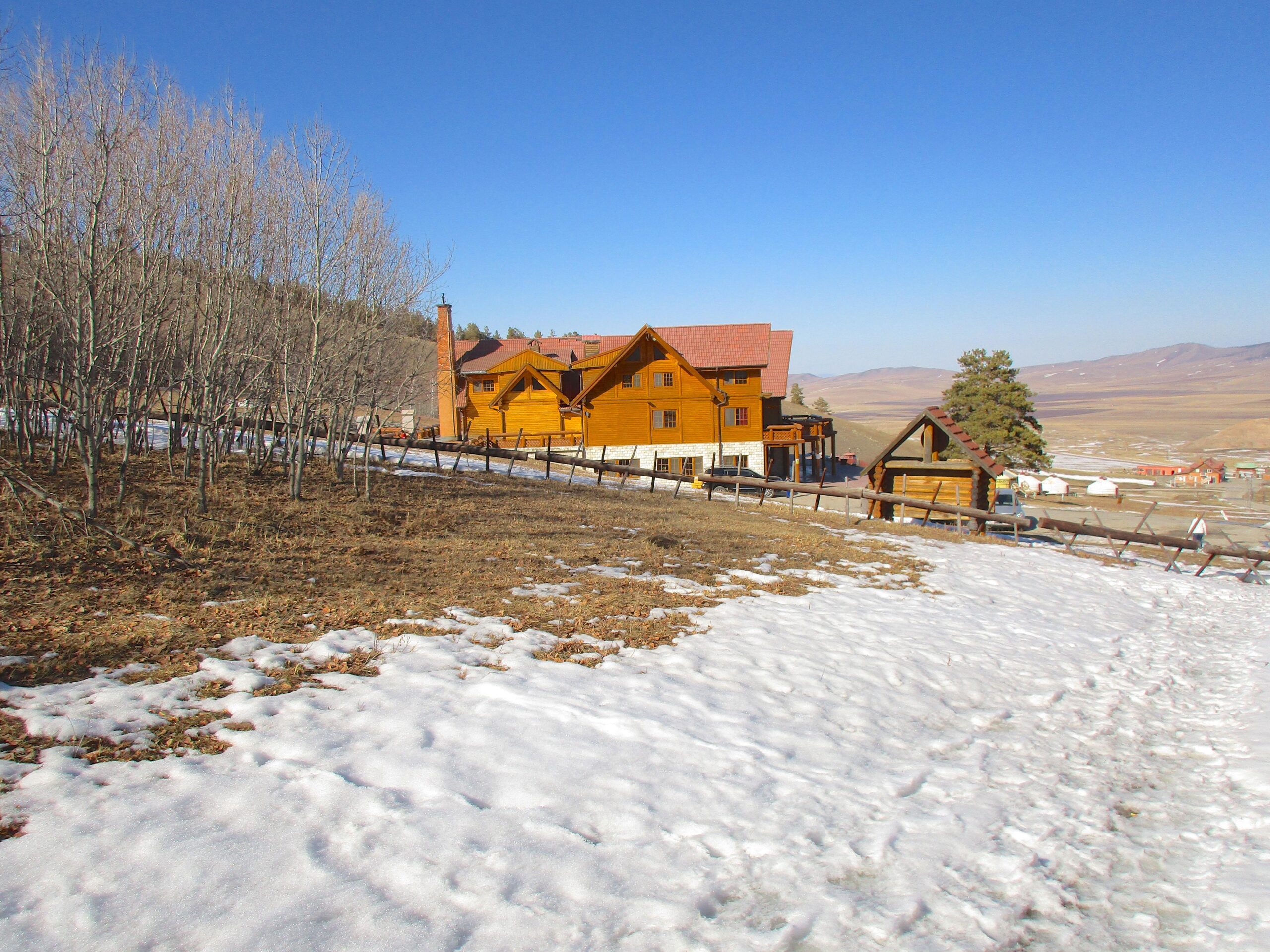 A scenic view of a wooden house surrounded by a snowy landscape and bare trees, with a clear blue sky in the background. The house features multiple stories and a red roof, while patches of snow can be seen beside grassy areas. In the distance, hills and a few small structures are visible, suggesting a tranquil, mountainous environment. Nuuts Tovchoo Amaralt mountain bike trail.
