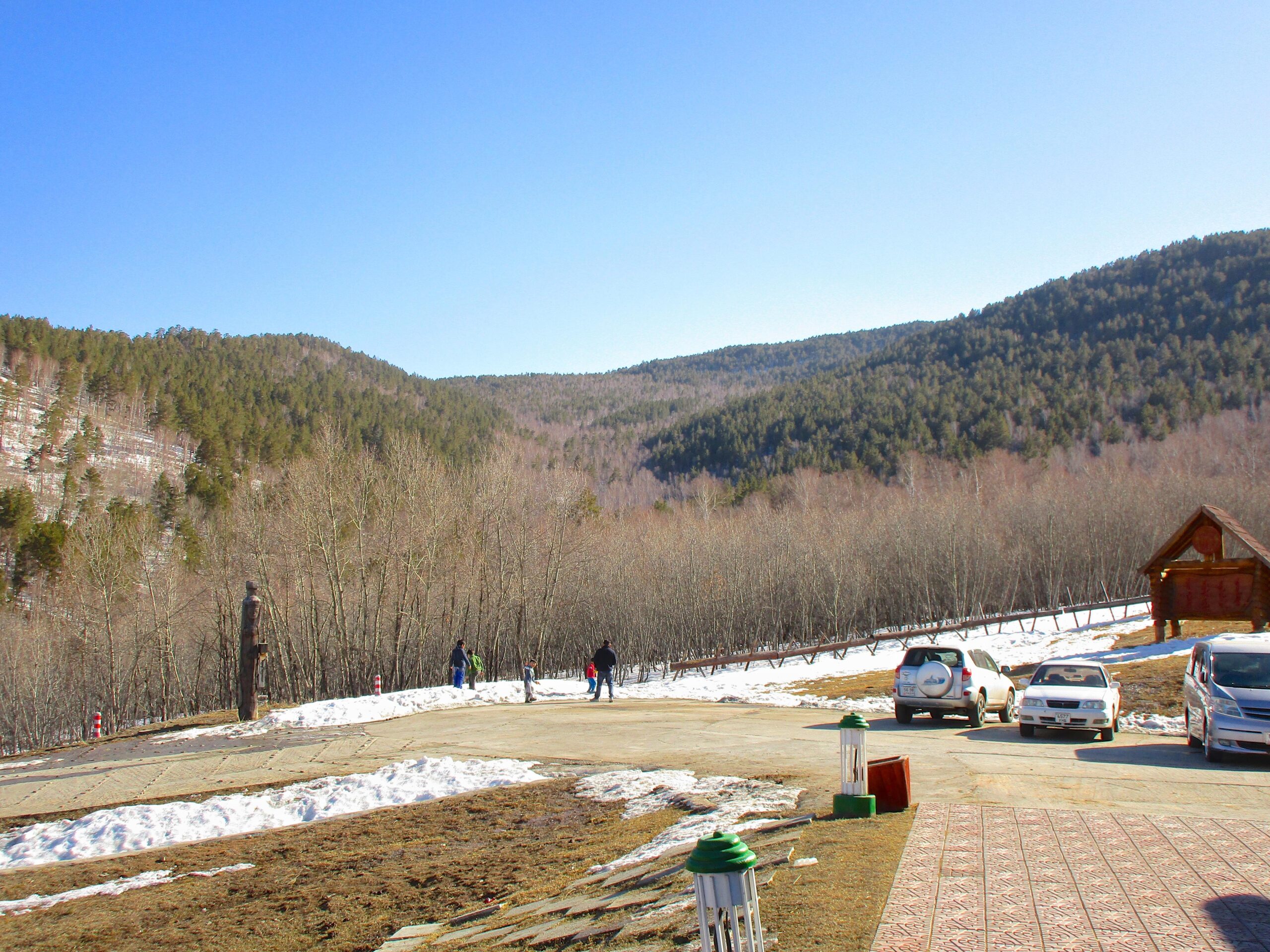 A scenic view of a mountainous landscape with a clear blue sky. In the foreground, there is a paved area with a few parked cars and several people walking, some carrying gear. Sparse patches of snow are visible on the ground, alongside a line of bare trees and green coniferous mountains in the background. A wooden structure is situated to the right, adding to the rustic charm of the setting. Nuuts Tovchoo Amaralt mountain bike trail.