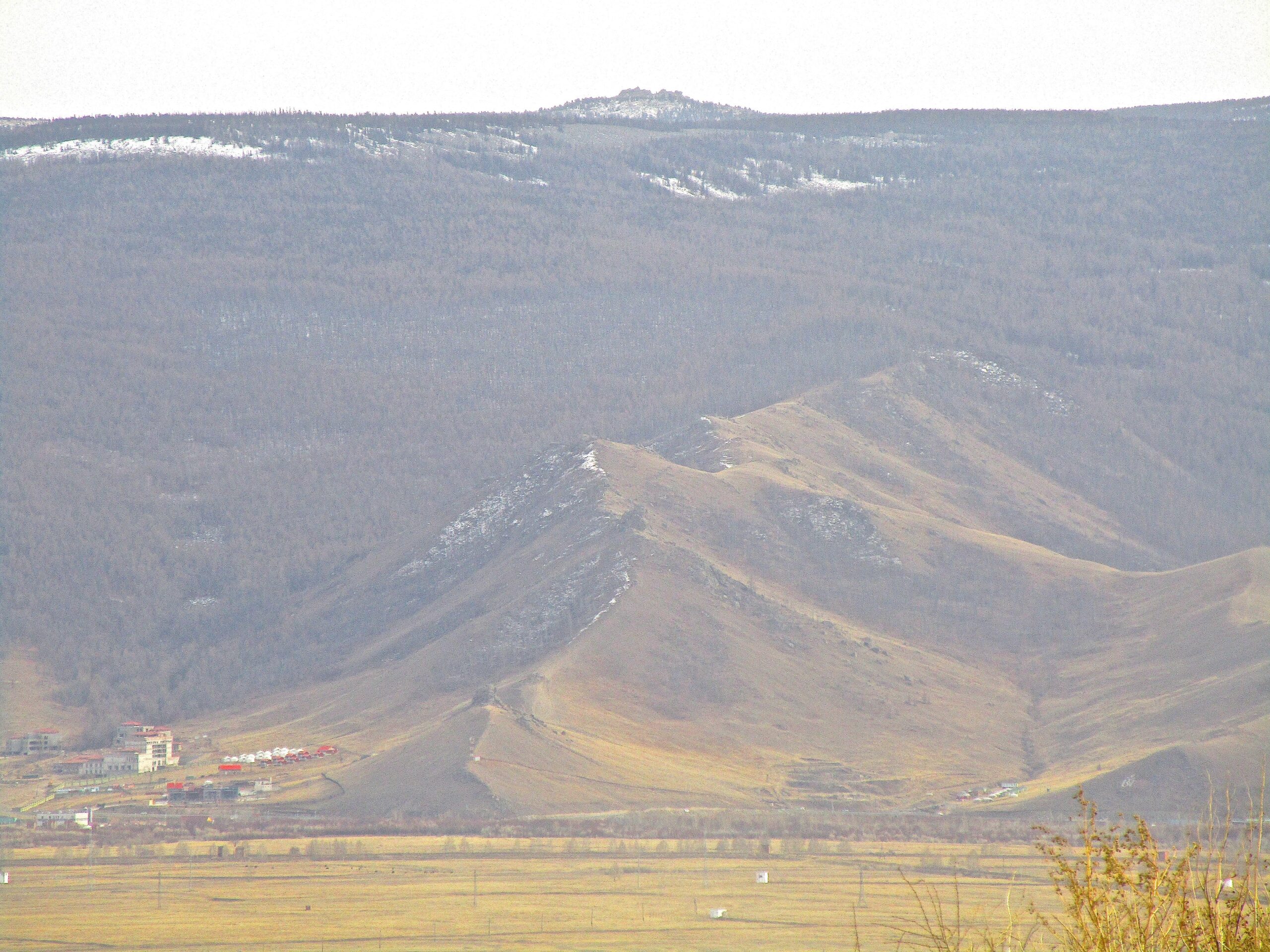 A mountainous landscape with rolling hills, green and brown vegetation, and a small settlement visible in the foreground. The hills lead up to a higher, snow-capped peak in the background under a cloudy sky. District 15 Network mountain bike trail.