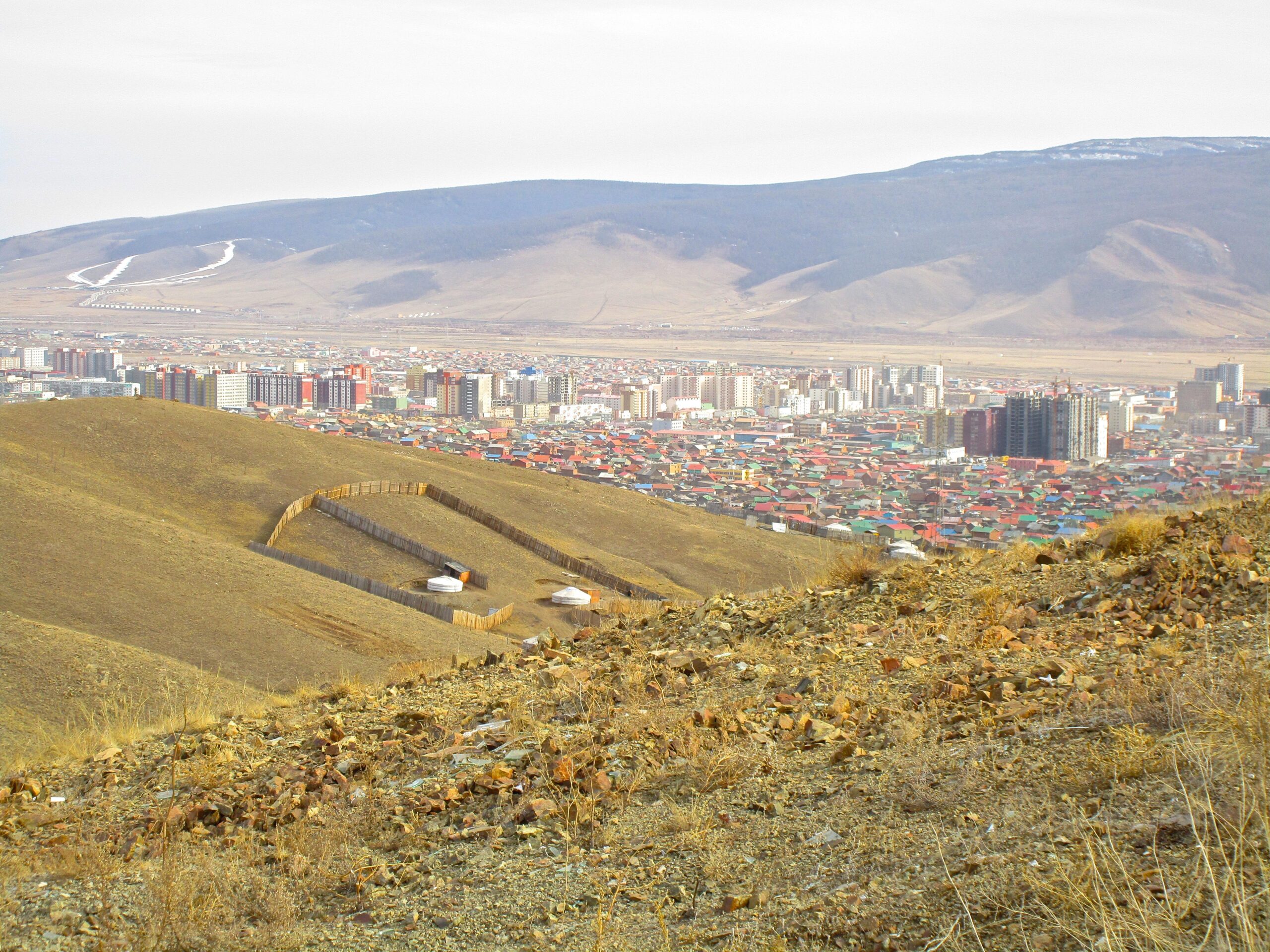 An expansive view of a city nestled in a valley, with colorful buildings and residential areas in the foreground. In the background, rolling hills are visible, and traditional yurts can be seen along a sloped hillside, suggesting a blend of modern and traditional architecture. District 15 Network mountain bike trail.